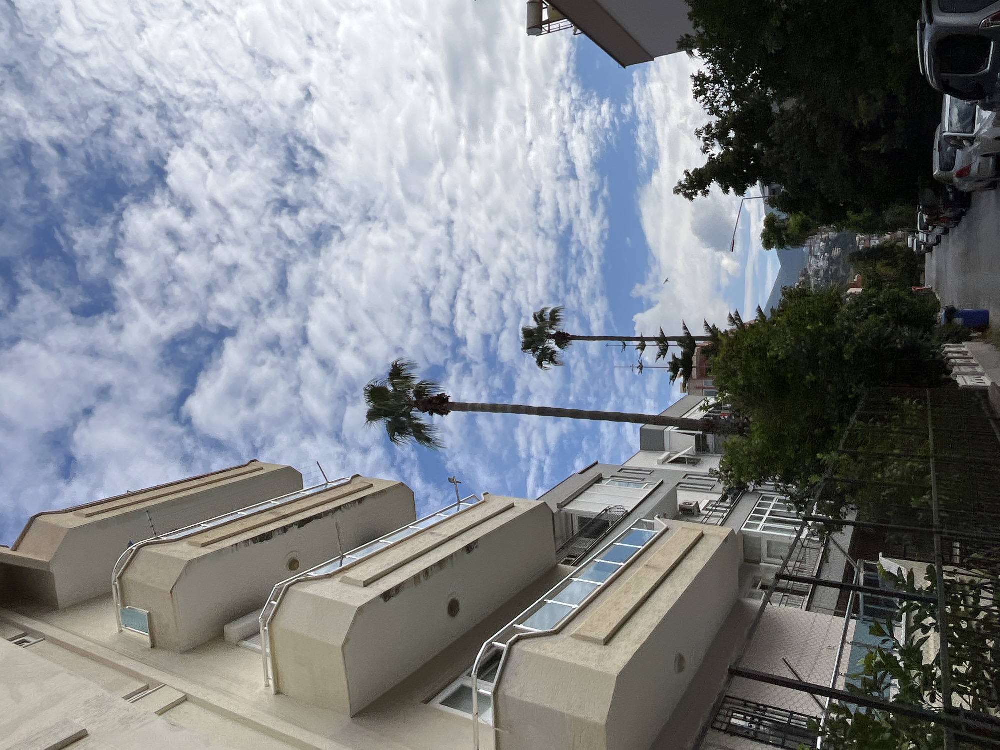 A low-angle, daytime outdoor shot in Alanya, Turkey. The frame is oriented vertically, capturing a perspective tilted upwards toward a sky filled with scattered white clouds. In the foreground, the architectural elements of a building are visible. These are light-colored, cantilevered structures extending from the building, possibly balconies or extensions. Railings and windows are integrated into these structures. The focus is on the buildings and the sky, with palm trees extending upwards. In the background, part of a neighborhood street can be seen. Parked cars line the side of the street adjacent to tree and some other houses.