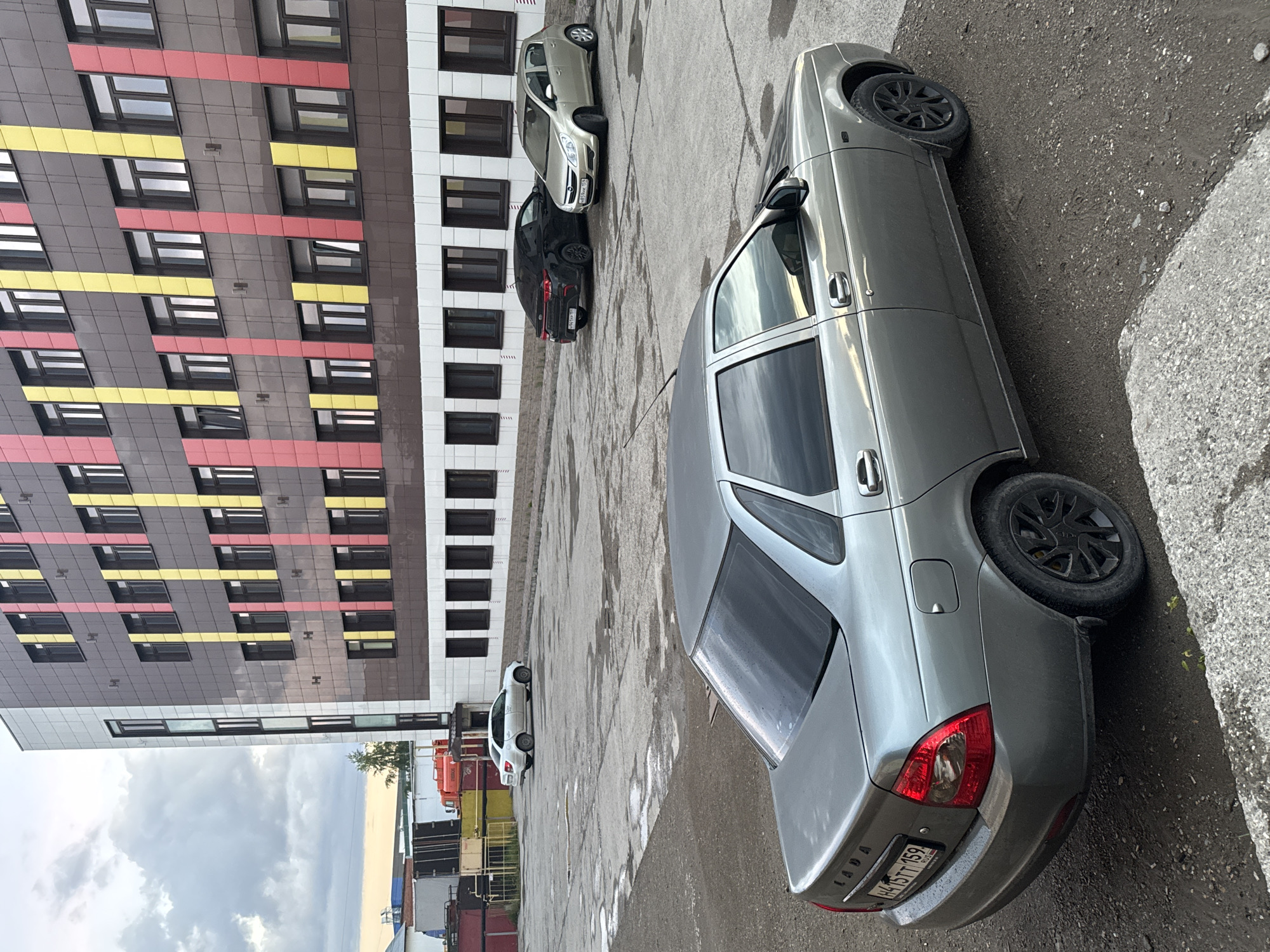 A grey Lada sedan is prominently parked on a wet, dark paved surface in what appears to be a parking lot or street in Perm, Russia. The car, featuring black alloy wheels, displays the Russian license plate "H416TT 159 RUS" on its rear bumper, with "LADA" also visible. The ground is damp, suggesting recent rain, with visible puddles and damp patches.
In the background, a modern multi-story residential building dominates the left side of the frame. Its facade is composed of dark grey panels interspersed with striking horizontal bands of yellow and red, creating a vibrant, geometric pattern. Numerous dark-framed windows punctuate the building's exterior. Adjacent to this building, another structure with a white facade and dark windows is partially visible.
Further along the paved area, several other vehicles are parked: a black sedan, a light-colored hatchback, and a white hatchback in the distance. The sky above is overcast with grey clouds, indicating a cloudy daytime setting. In the far distance, beyond the parking area, some industrial containers or structures can be discerned. The overall scene depicts a typical urban environment with modern architecture and parked vehicles under a cloudy sky.