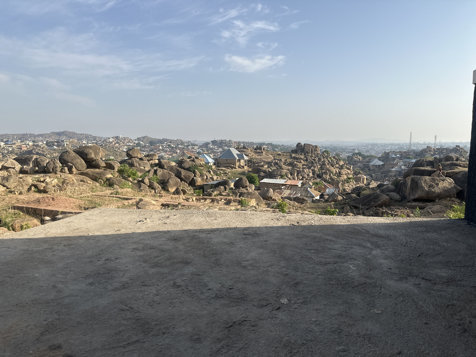 The photograph depicts a rocky landscape with scattered buildings in Jos, Nigeria. In the foreground is a concrete platform or floor. Beyond this platform, the terrain is dominated by large, smooth boulders interspersed with sparse vegetation and buildings with corrugated metal roofs. In the distance, the urban sprawl of Jos is visible under a hazy, blue sky with wispy clouds. One person is visible on the right side of the frame, standing on a large rock.
