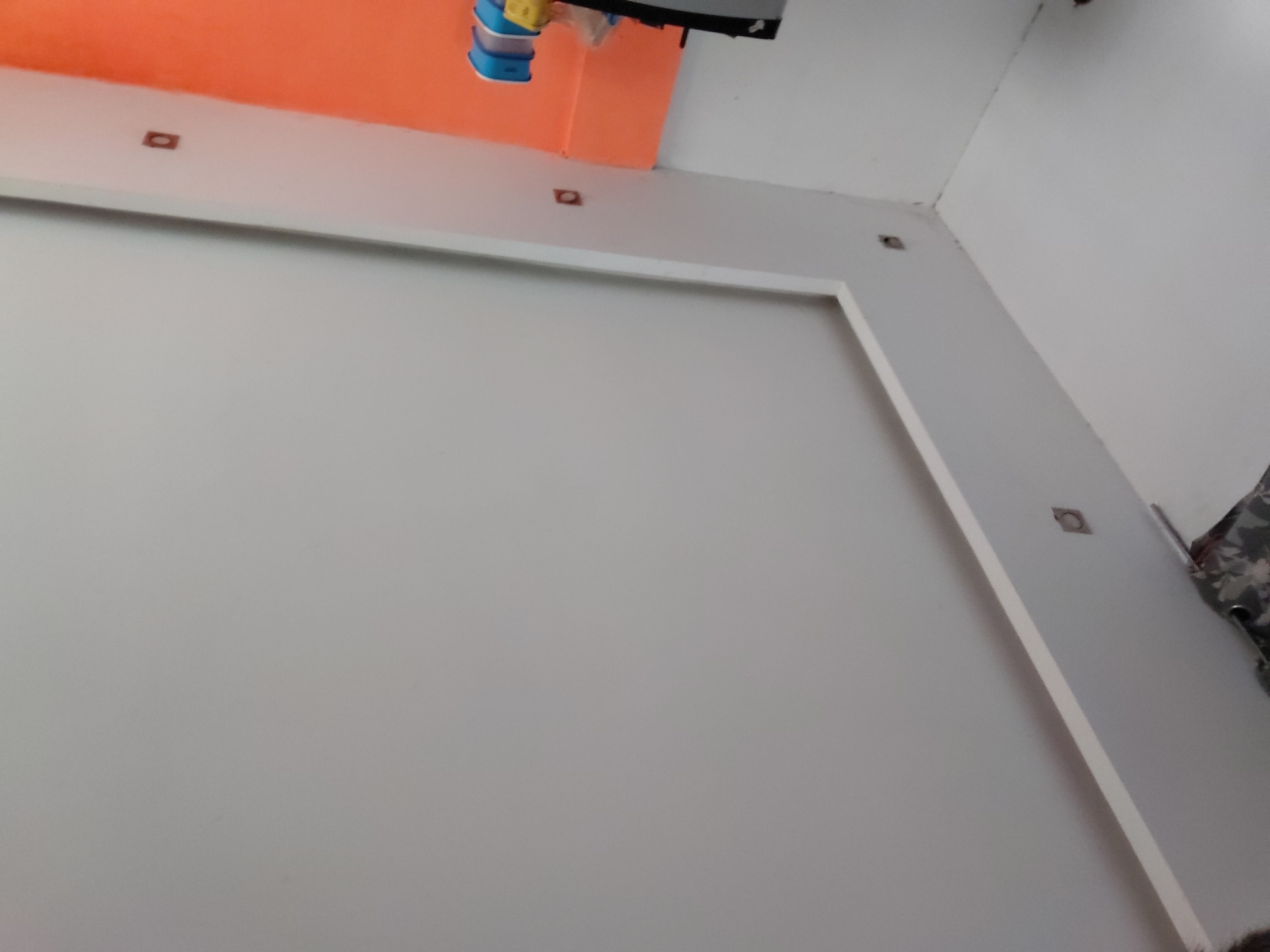 The image depicts an interior architectural space, captured from a tilted, low-angle perspective. The primary subject is a large, light grey wall, which features a prominent, rectangular or square frame-like structure in a lighter, possibly white, material. This frame creates a decorative or recessed panel effect on the wall.
Above the grey wall, an upper section or ceiling transitions, painted in a distinct bright orange color. Several small, dark, square or rectangular fixtures are embedded into the ceiling and the white wall section to the right. These could be recessed lighting units or mounting points for other elements.
In the upper left corner, a small object with blue and yellow components is visible, appearing to hang from an unseen fixture above the frame. On the far right side of the image, a piece of dark fabric with an indistinct light pattern is partially visible. A small, dark, fuzzy shape at the very bottom right corner might be a person's hair or part of an object on the floor.
No specific activity or event is taking place, and no individuals are clearly visible. The lighting appears even, suggesting an indoor setting under artificial or diffused natural light. There is no visible text in the image. The scene portrays a modern or minimalist interior design in Santa Ana, Venezuela.