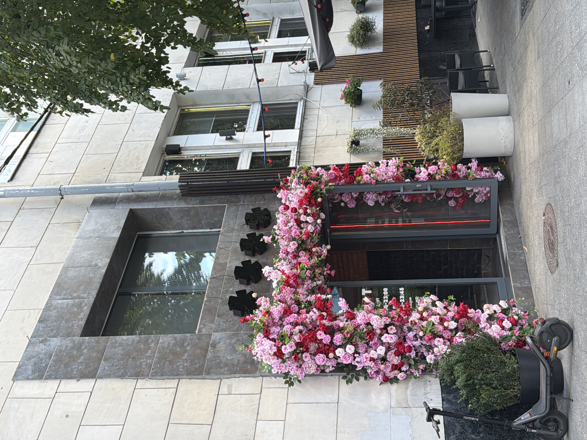 An exterior shot captures the entrance to a building adorned with a vibrant floral arch. The building's facade is constructed of large, light-colored stone tiles. Above the entrance, a dark grey rectangular frame surrounds a window reflecting trees and the sky. The entrance itself is a doorway framed by an abundance of pink and red flowers, creating a lush archway. Black, star-shaped motifs are visible above the floral arrangement. To the left of the entrance, a black electric scooter is parked next to a large potted green plant. To the right of the entrance, the scene opens up to a patio area with wooden slat walls, outdoor seating with dark chairs and tables, and additional potted plants. Several windows with decorative lighting are visible on the upper floors of the building. The ground is paved with light grey tiles. A tree with green leaves is visible on the right side of the image. No people are present in the scene. The overall impression is of a stylish establishment with a welcoming, floral-themed entrance.