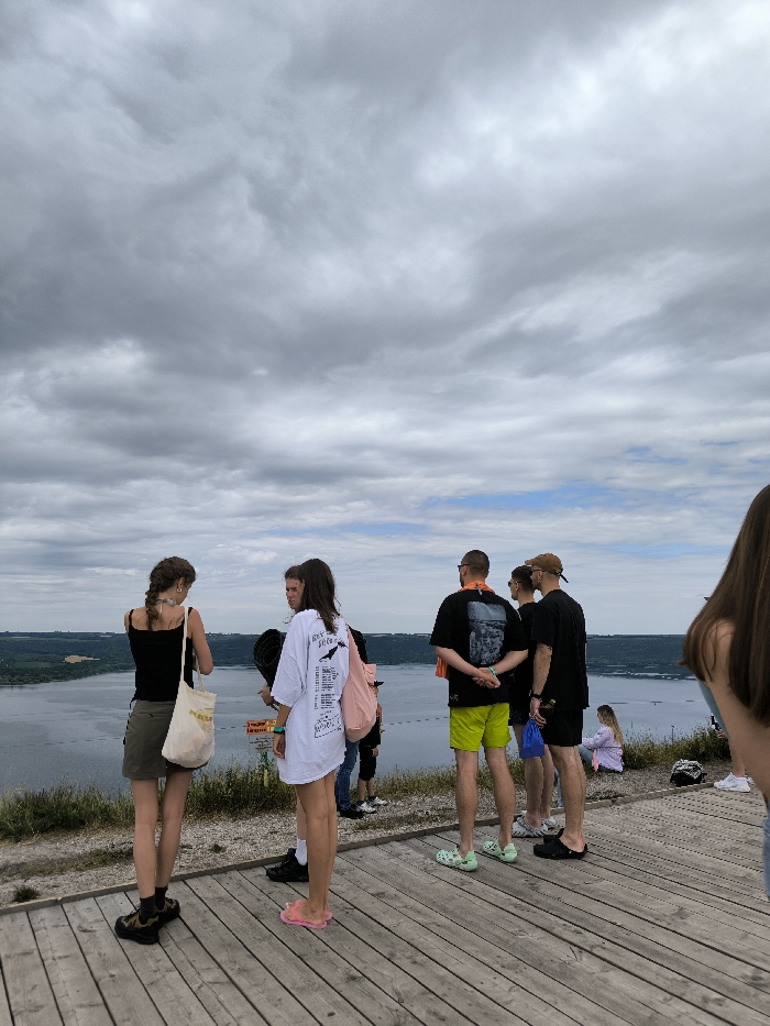 The image depicts a scenic overlook in the Horaivka area of Ukraine, providing a panoramic view over the Dniester River. The sky is largely overcast with varying shades of grey clouds, though hints of blue are visible towards the distant horizon above the river. The wide Dniester River flows through the midground, its banks lined with dense green vegetation.
In the foreground, a light-colored wooden deck or platform serves as the viewing area. Nine individuals are present on or near this platform. On the wooden deck, a group of six stands, primarily facing towards the river. This group includes two females: one in a black tank top, an olive-green skirt, and carrying a light tote bag; the other in an oversized white t-shirt with black graphics, pink flip-flops, and a pink backpack. Among the males in this group, one wears bright green shorts and light green clogs, another a light brown cap. To the immediate left and far right, two additional individuals are partially visible. Further back and to the right, on a grassy slope, one person sits on the ground facing away from the camera. The individuals appear to be observing the natural landscape.
