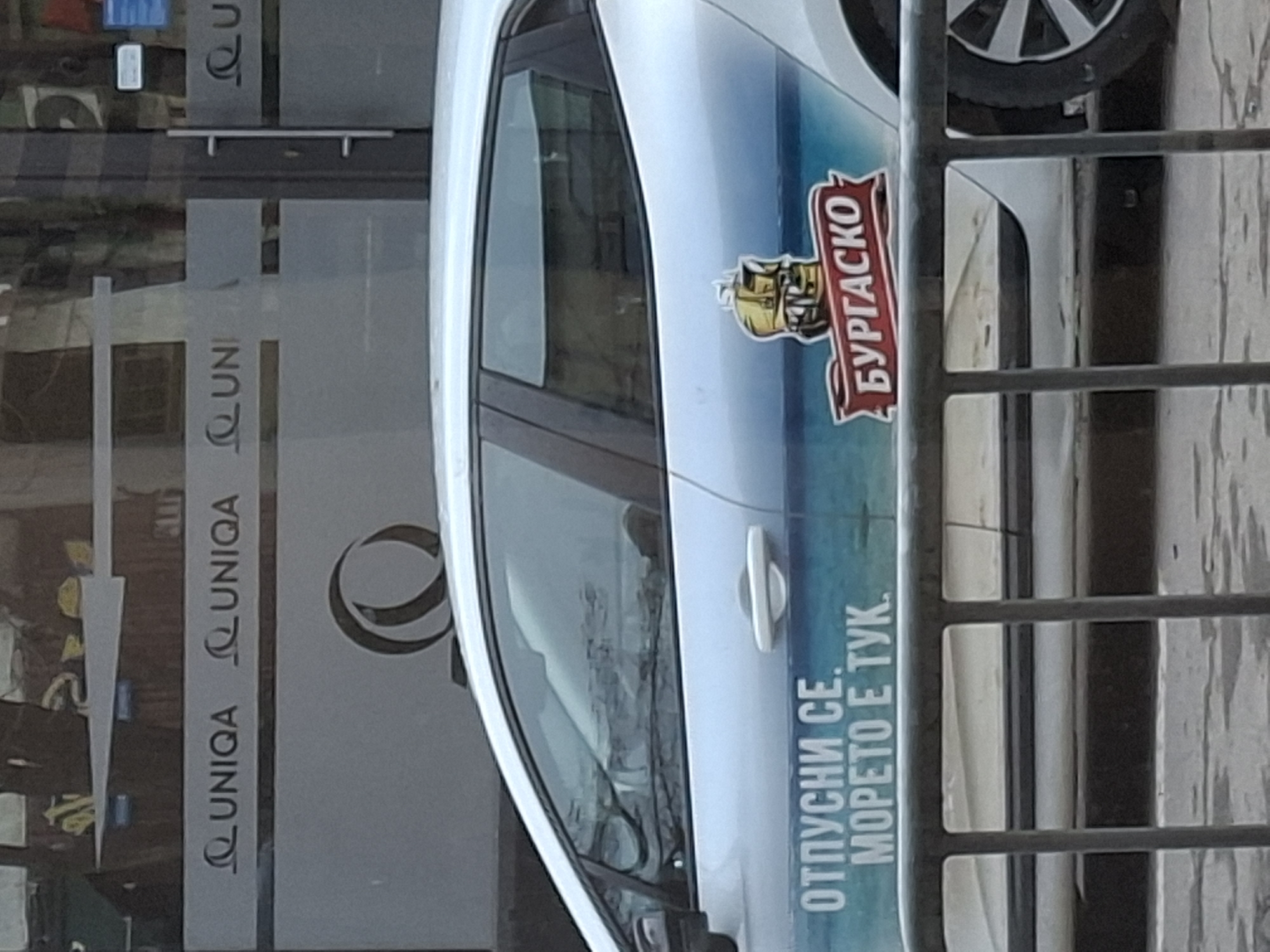 The image shows a partial view of a white car parked near a UNIQA insurance office in Sofia, Bulgaria. The car has an advertisement for "Burgasko" beer, featuring a ship and the phrase "Отпусни се. Морето е тук." (Relax. The sea is here.). A metal railing is partially obscuring the view of the car.