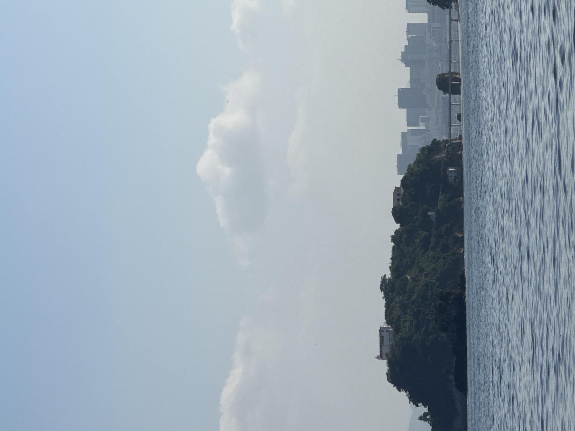 The image shows a view of the city of Niterói, Brazil, from across the water. In the foreground, the blue water is rippled. In the middle ground, a green, tree-covered island with buildings on top sits in the water. A bridge spans across the water from the island to the mainland. In the background, a hazy cityscape with tall buildings is visible. The sky is light blue with scattered white clouds.