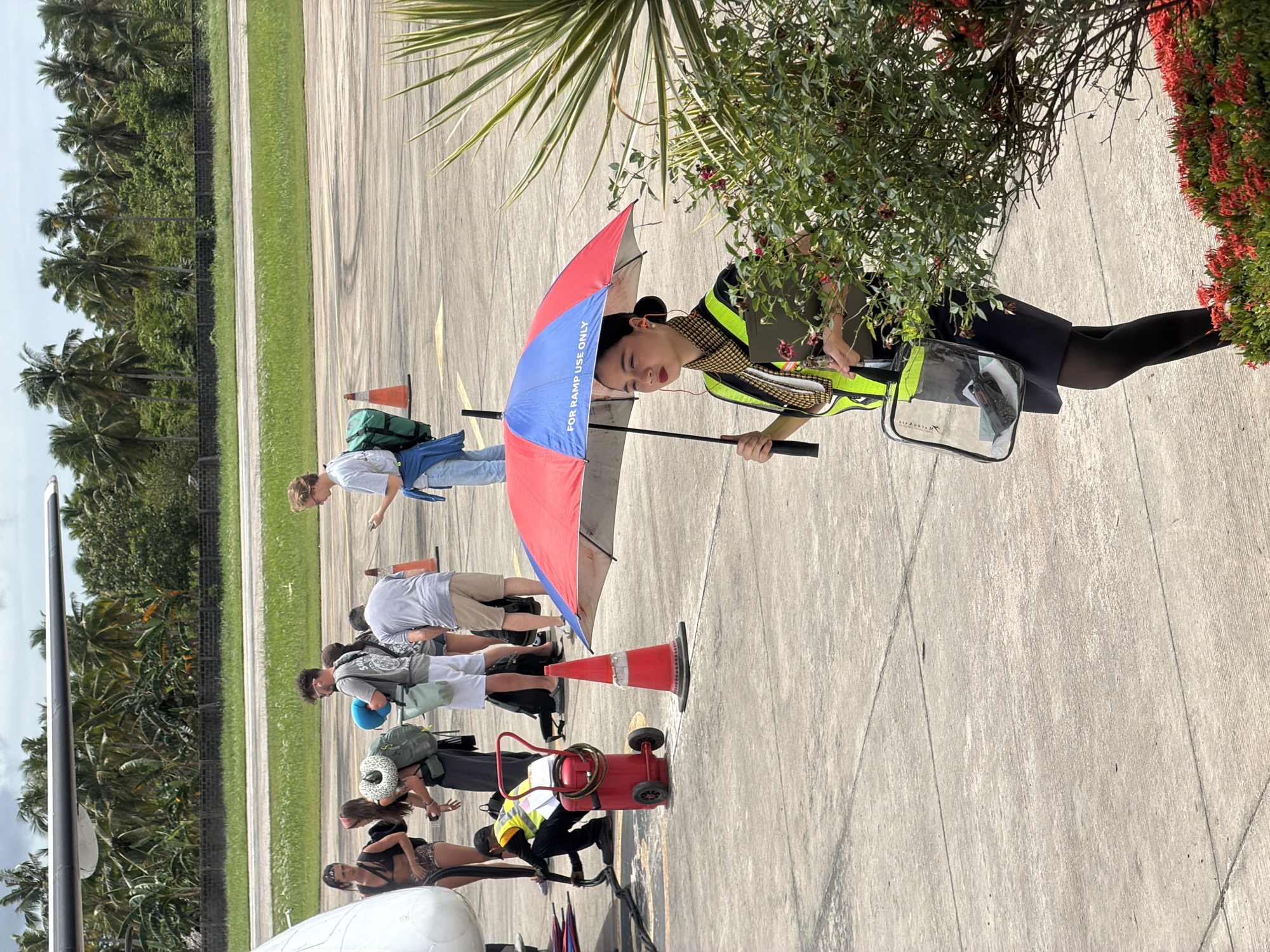 The scene appears to be at the Del Carmen Airport in the Philippines. A line of passengers disembarks an aircraft on the tarmac. An airport employee wearing a high-visibility vest holds a blue and red umbrella marked "FOR RAMP USE ONLY" and a clear bag. There are traffic cones, luggage, and a small cart near the aircraft. Tropical vegetation surrounds the tarmac area.