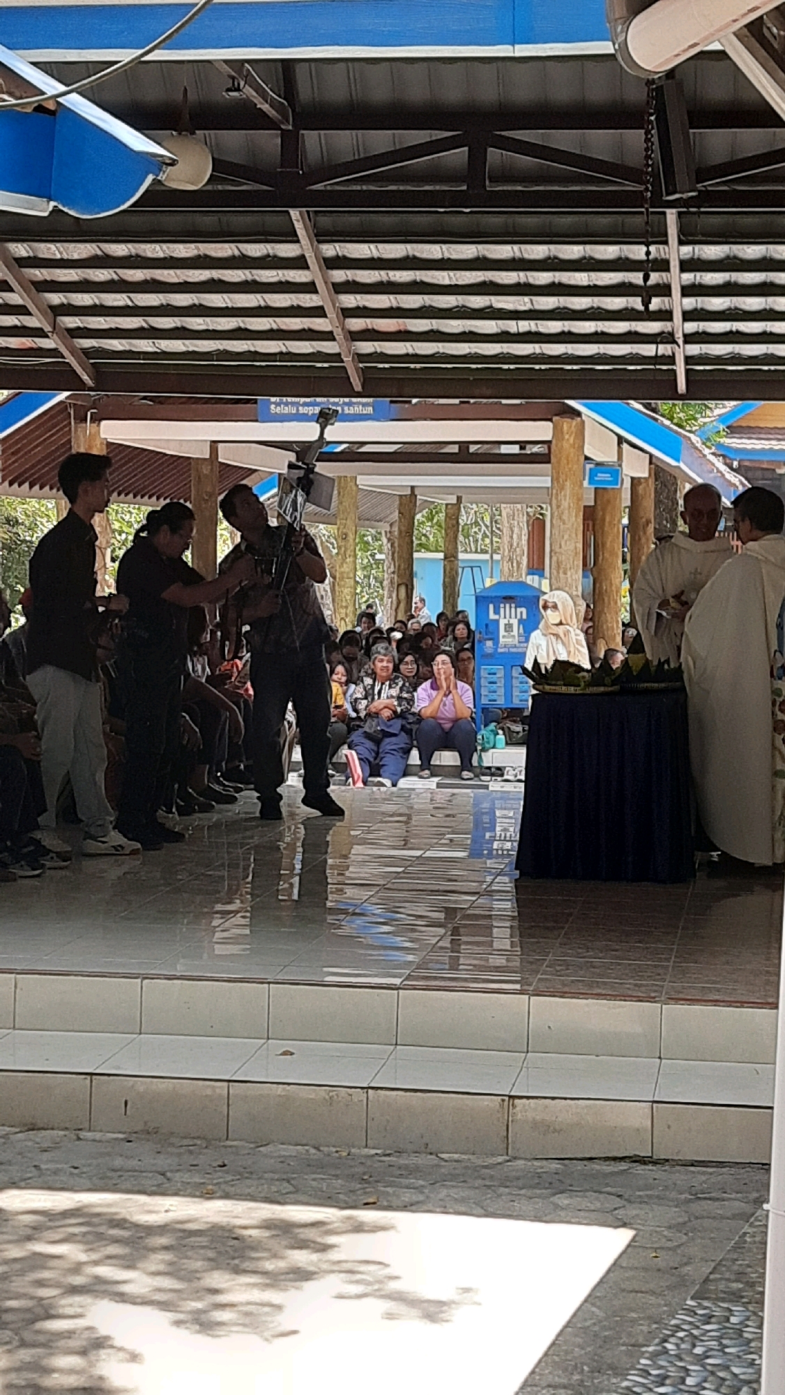 In Moyudan, Indonesia, at a tiled outdoor venue with a partial roof structure, a ceremony is underway. Three individuals are prominent in the foreground: a person on the left in a black shirt and light-colored pants, holding a camera; another person in a black shirt in the center, adjusting a camera on a tripod; and a third person on the right, a clergyman in a white robe, interacting with another clergyman also in a white robe. A dark blue tablecloth covers a table on which sits a sculpture and some decorative green foliage. Spectators are seated behind the table, some looking towards the clergymen. A blue sign with white text is visible in the background. The floor is highly reflective, creating a mirror-like effect.