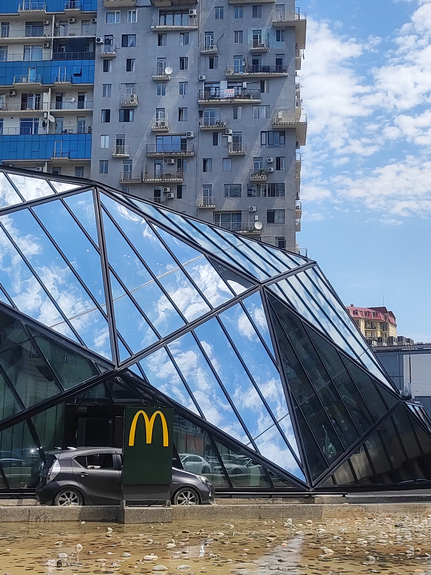 The image shows a modern McDonald's restaurant with a distinctive geometric glass facade located in Batumi, Georgia. A dark gray car is parked in front of the restaurant. The McDonald's logo, a golden "M" on a green background, is prominently displayed. In the background, a tall, gray apartment building with blue accents and multiple balconies is visible, along with another building with a red roof. The sky is blue with scattered white clouds, and the foreground features a shallow pool of water with scattered stones.