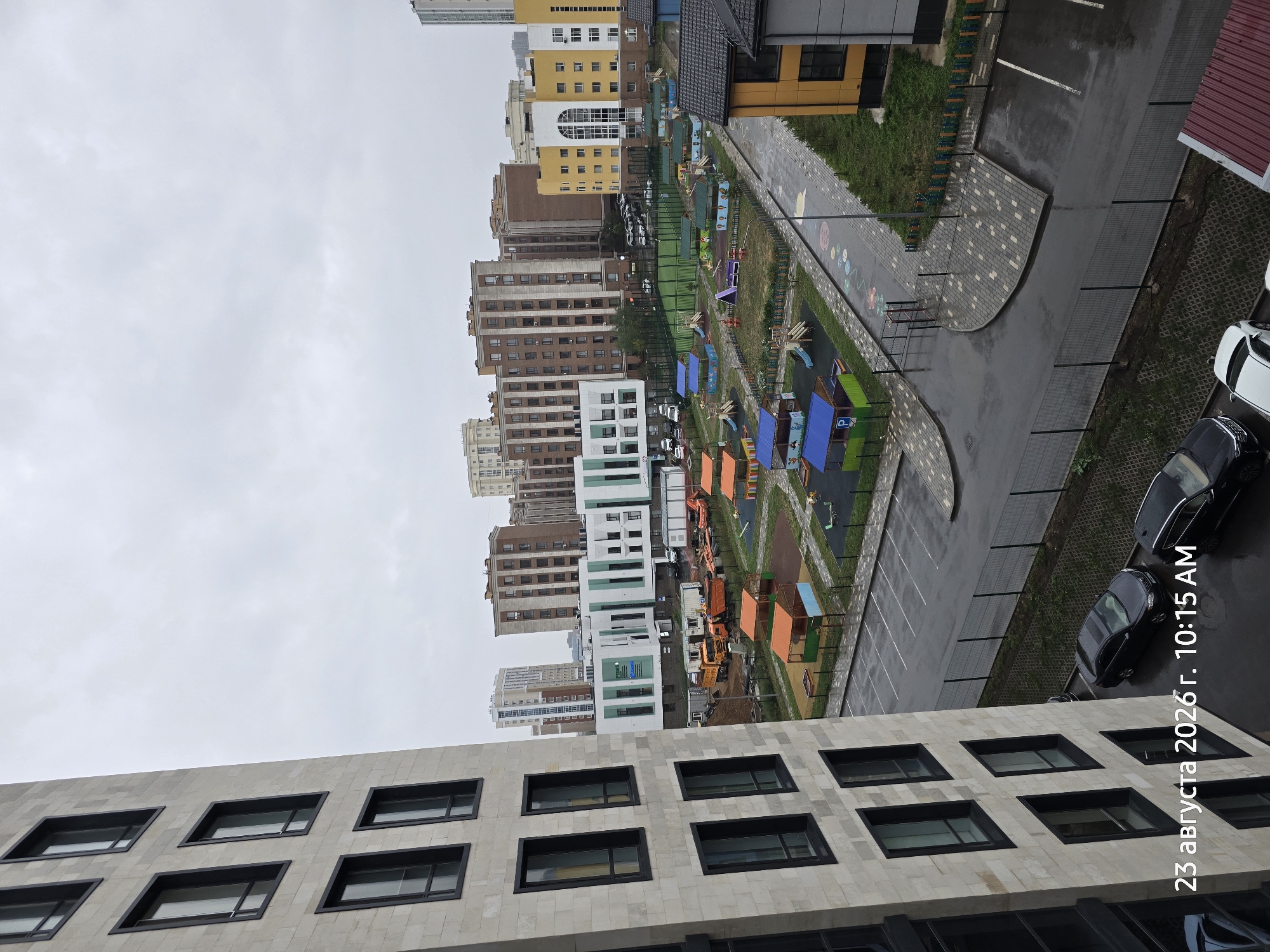This is a high-angle, eye-level, full shot of a residential area in Astana, Kazakhstan, on a cloudy day. The timestamp "23 августа 2026 г. 10:15 AM" is overlaid on the bottom left of the image. In the foreground, a modern, light-colored residential building with multiple floors and dark-framed windows is visible on the left. To the right of this building, across a wet paved area with parking spaces, is a children's playground with various colorful structures including slides, canopies, and sandboxes. Beyond the playground are several multi-story apartment buildings of varying architectural styles and colors, including shades of brown, beige, and yellow. Some of these buildings appear to be under construction or recently completed, with visible construction equipment like trucks and containers in the middle ground. The sky is overcast and gray. Several cars are parked in the paved areas and on the street. The ground in the playground and parking areas is wet, suggesting recent rain.
