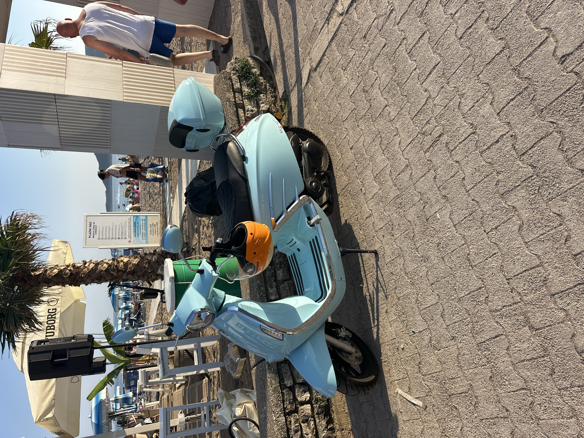 An eye-level medium shot captures a light blue scooter parked on a paved walkway near a beach in Boreti, Montenegro. The scooter is positioned facing left, with its kickstand down. A bright orange helmet with a black visor is attached to the handlebars. Behind the scooter, a stone wall runs along the edge of the walkway, topped with green plants. To the left of the scooter, a white outdoor seating area with tables and chairs is visible, partially obscured by a large white umbrella bearing the "Uborg" logo. A tall black speaker stands behind the umbrella, next to a palm tree. In the background, beach umbrellas and loungers suggest a coastal setting, and people can be seen walking on the sand and in the water. The paved surface consists of interlocking brick-like stones. A person wearing a white tank top and blue shorts walks away from the viewer on the right side of the frame, near a large, textured column.