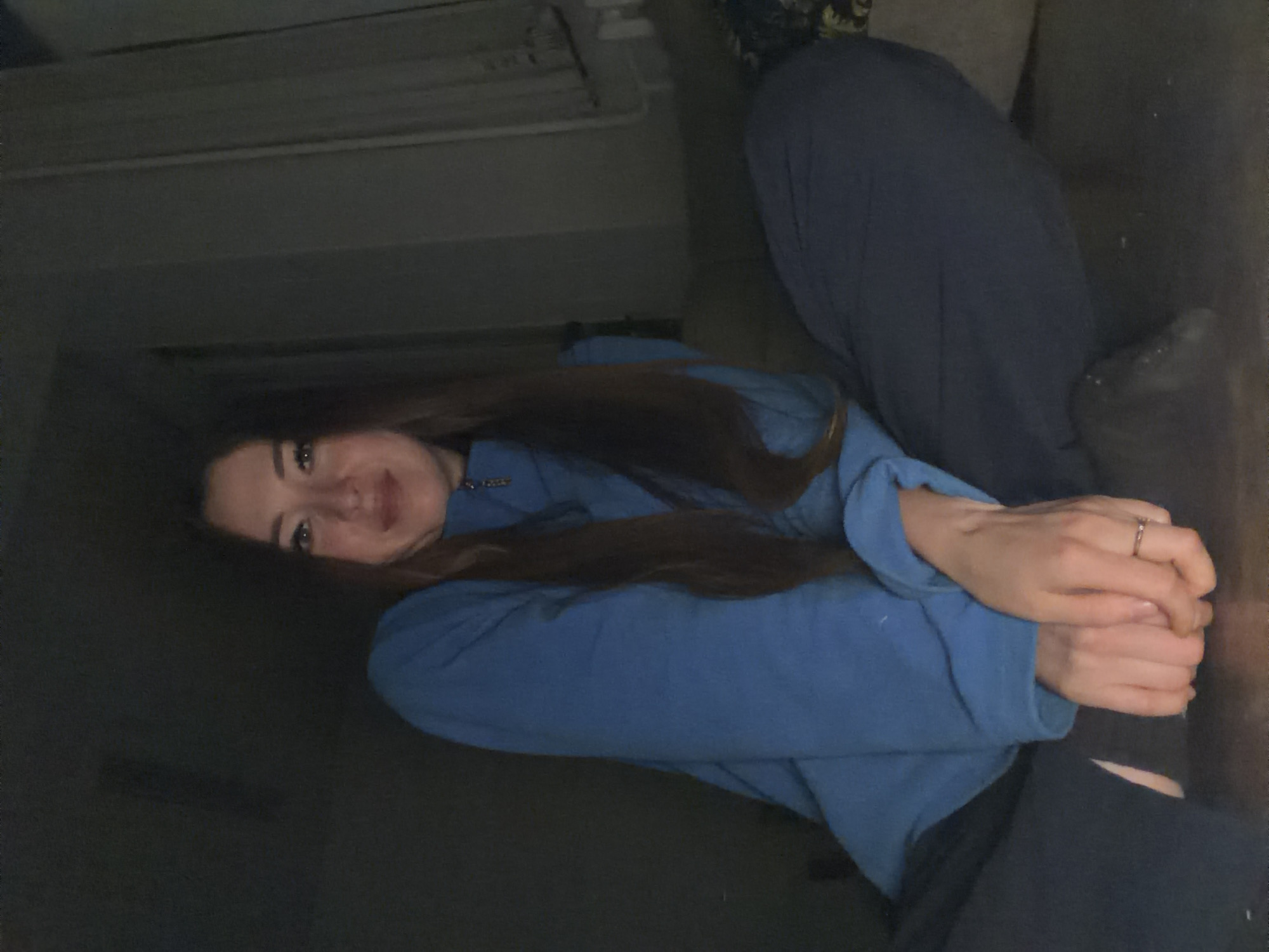 The image shows a young woman in a dimly lit room, possibly a living room or bedroom, based on the presence of a couch and what appears to be a radiator or air conditioning unit on the wall in the background. The woman has long, dark brown hair and is wearing a blue sweatshirt. Her hands are clasped together in front of her, and she is wearing a ring on her right hand. She is looking towards the camera with a slight smile. The overall lighting is low, suggesting it might be nighttime or a dimly lit interior. There are no visible signs of weather conditions or specific time of day beyond the lighting. The setting is domestic. There is no visible text in the image.