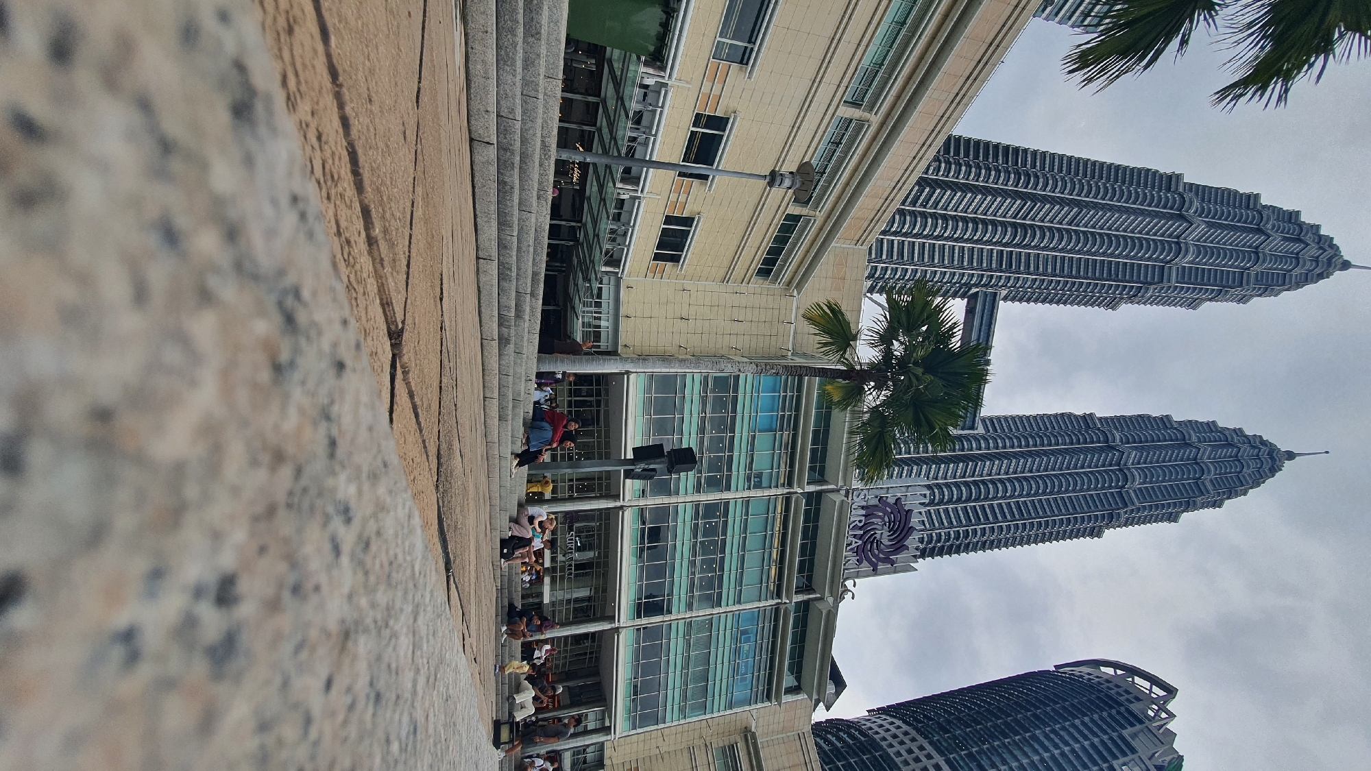 The image depicts the Petronas Twin Towers in Kuala Lumpur, Malaysia, viewed from a low angle. The foreground features a textured stone surface. Behind it, a wide set of stone steps leads up to a modern building complex, identified by a sign as "Suria KLCC." Several people are seated and standing on the steps, appearing relaxed. Palm trees are visible, with one prominent tree positioned between the building and the towers. The Petronas Twin Towers, iconic skyscrapers, dominate the background, connected by a skybridge. The sky is overcast with a cloudy, grayish hue. The scene appears to be an outdoor public space, likely a plaza or park area adjacent to the shopping mall at the base of the towers.