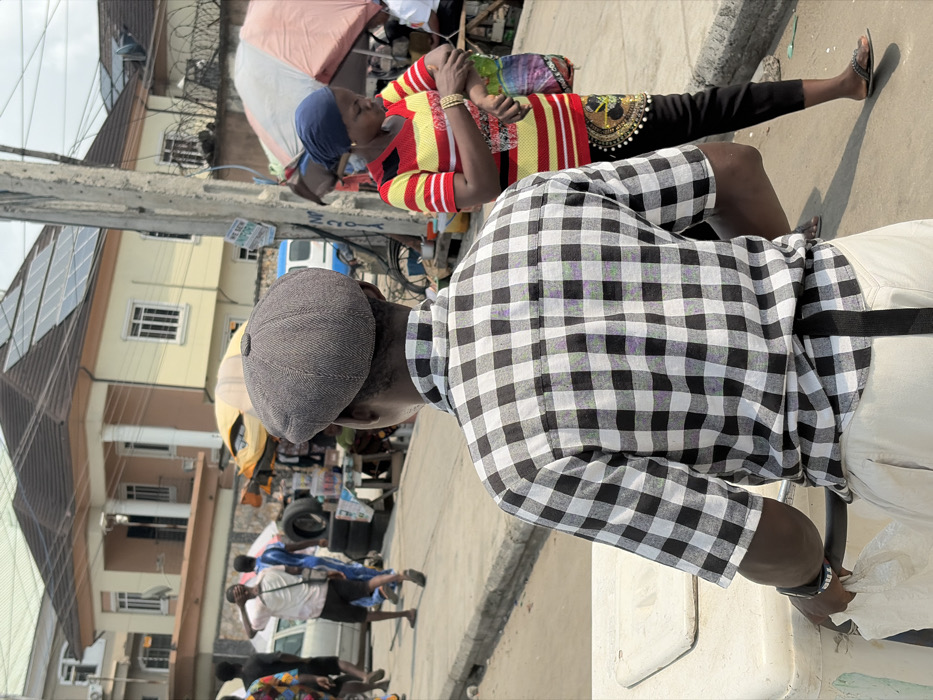 The scene depicts a bustling street in Jakande, Nigeria, on a clear day. In the immediate foreground, a dark-skinned male is viewed from behind, wearing a grey herringbone flat cap, a short-sleeved black and white plaid shirt, and light-colored trousers with a black strap hanging from the waist. His left wrist bears a black watch, and he grips a light-colored, rectangular object, possibly a cooler or vendor's cart, at the bottom left.
To the right of the foreground subject, a dark-skinned female, seen from the waist up, wears a dark blue headscarf, a red and yellow striped short-sleeved shirt, and black pants. Gold-colored bangles adorn her left wrist as she holds a colorful patterned plastic bag in her left hand, her right hand near her left forearm. She wears black sandals.
The midground features a concrete curb separating the subjects from a street populated by pedestrians and roadside activity. A tall, grey utility pole stands centrally, covered in wires and bearing a faded sign. Behind the female, a large, light-colored umbrella with red/orange stripes provides shade. Further down the street, multiple individuals are visible, including a man in a white t-shirt scratching his head, and a light-colored sedan parked on the left. Several makeshift stalls display goods, including stacks of tires and various merchandise under a yellow awning.
The background is dominated by a two-story, pale yellow building with a dark, sloped roof fitted with multiple solar panels. White columns and numerous windows are visible on the building's facade. Another building is partially visible to the right. The sky appears bright and somewhat hazy.