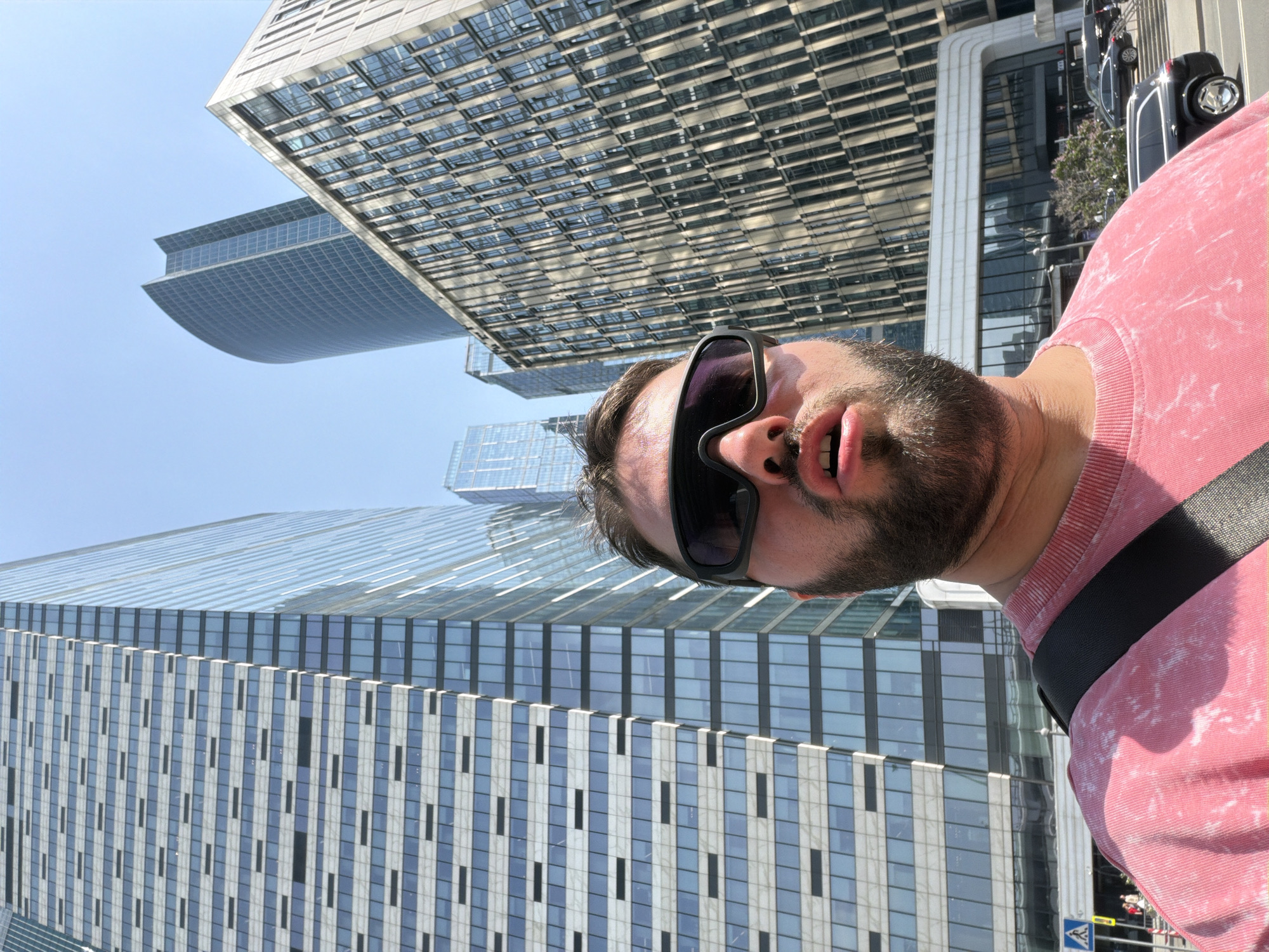 A man wearing sunglasses and a pink shirt is posing for a selfie. In the background are several modern high-rise buildings with glass facades and metal or concrete structures. Some vehicles are also visible. The location is Khimki, Russia.
