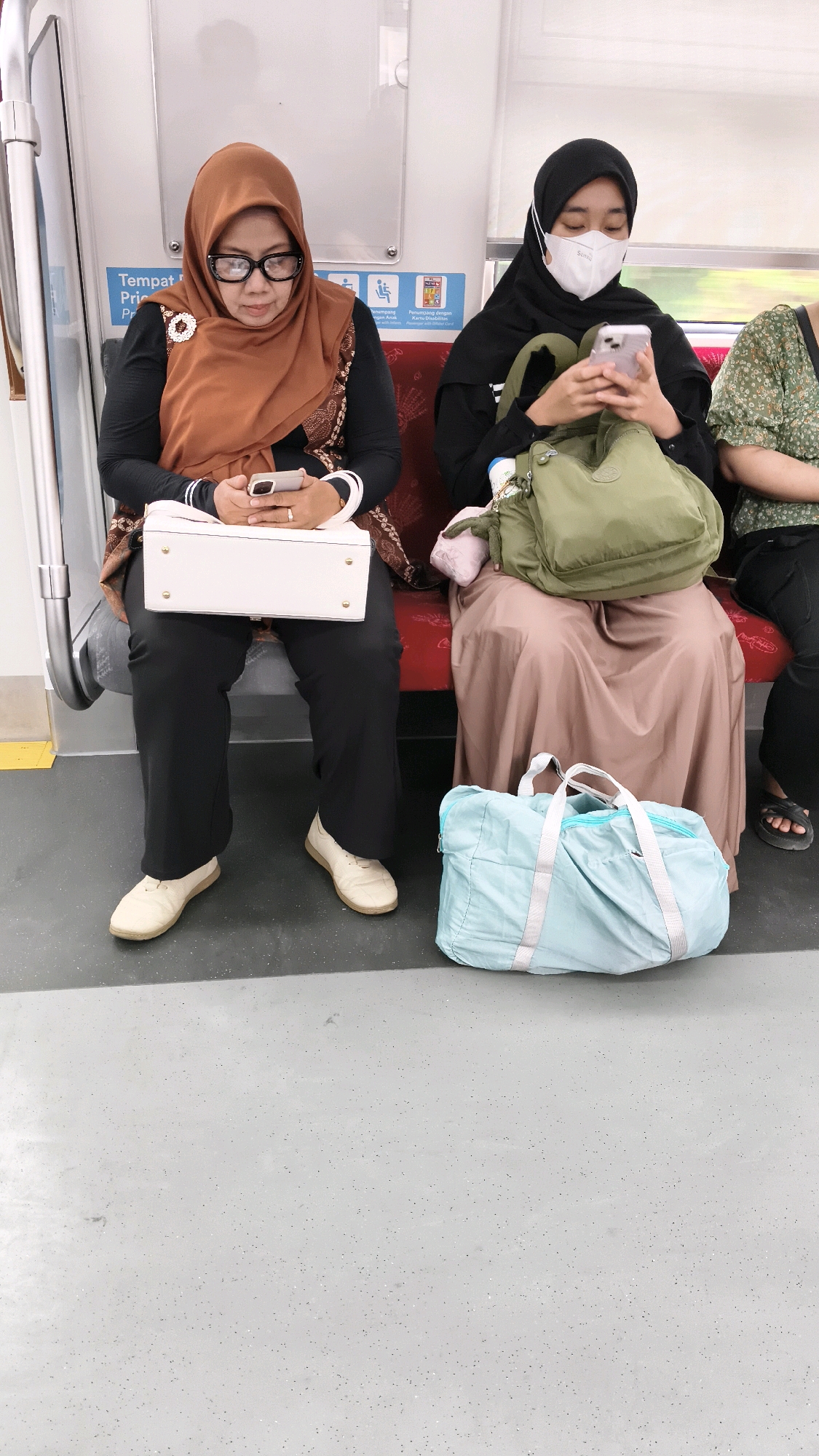 In Bekasi, Indonesia, three women are seated on a train. The woman on the left wears a brown hijab and glasses, holding a white purse and a smartphone. The woman in the middle wears a black hijab and a white face mask, also holding a smartphone and a green bag. The woman on the right is partially visible, wearing a green patterned top. Two bags, one white and one light blue, are on the floor in front of them. The train's interior features red seats and a gray floor.