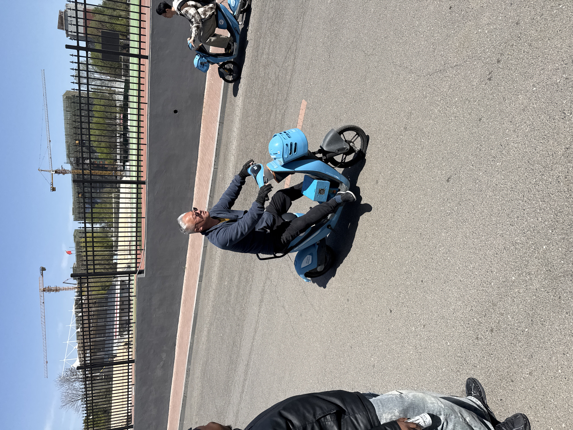 The image shows an outdoor scene in Zhongtun, China, featuring a man sitting on a blue electric scooter on an asphalt surface. He is wearing sunglasses, a dark jacket, and black pants. Another person is also riding a similar blue scooter in the background. In the foreground, a portion of a person wearing a black jacket and jeans is visible. A black metal fence lines the side of the road, behind which a building under construction with visible cranes can be seen. The sky is clear and blue.