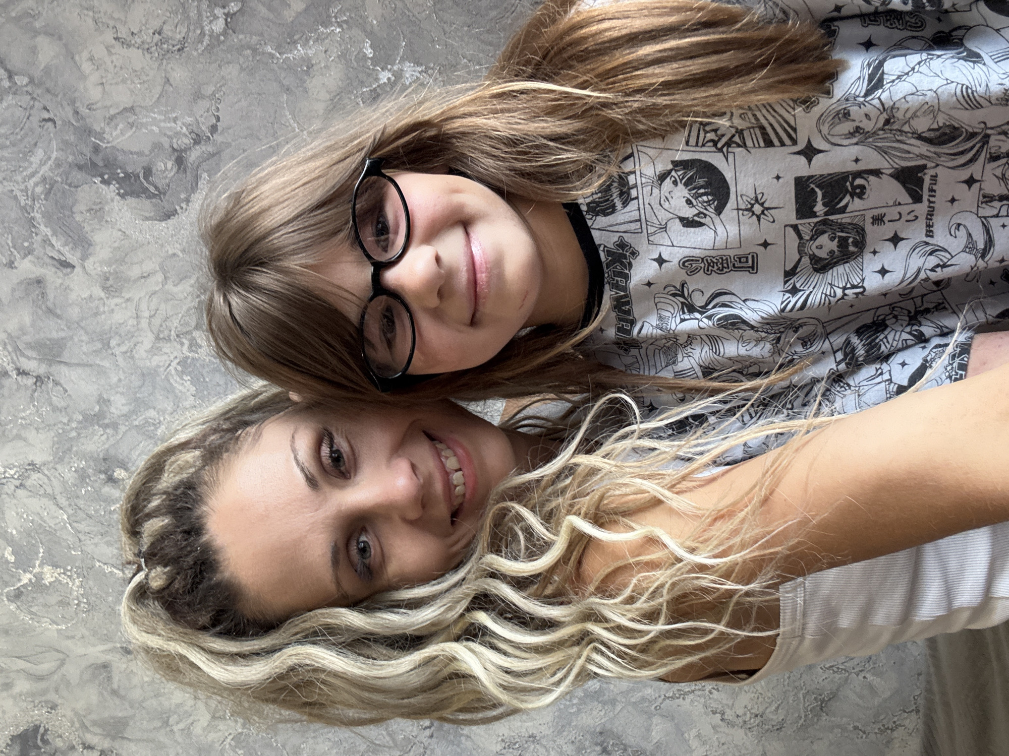 In an indoor setting with a textured gray wall as the background, a woman and a young girl are captured in a close-up, shoulder-up portrait. The woman, positioned on the left and slightly behind the girl, has blonde, wavy hair pulled back into a high ponytail and is wearing a white tank top. She has a warm smile, revealing slightly spaced front teeth, and is looking directly at the camera. Her right arm is around the girl. The girl, on the right, has long, straight brown hair and is wearing glasses with black frames. She is also smiling, with dimples visible on her cheeks, and is looking forward. She is wearing a light-colored t-shirt with a black and white pattern of anime-style illustrations and text. The lighting appears to be natural, creating soft shadows and highlighting the textures of the background and the subjects' features. The location is stated to be in the city of Rusanovo, Russia.