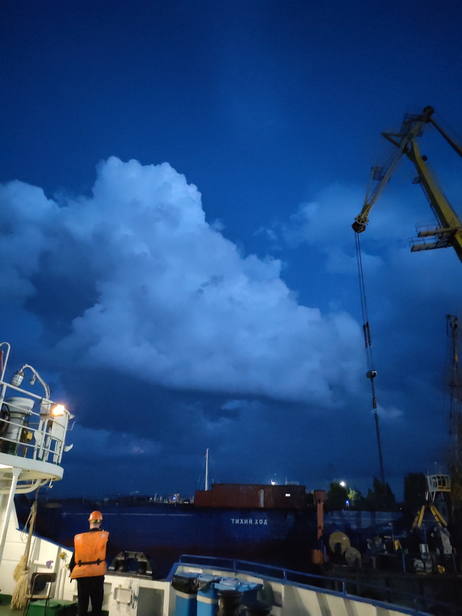 The image depicts a port scene at dusk in Rybinsk, Russia. A person wearing an orange life vest and hard hat stands on the deck of a white vessel, looking towards a large blue ship. The blue ship has Cyrillic lettering that reads "ТИХИЙ ХОД" (Tikhiy Khod, meaning "Quiet Passage"). A yellow crane is visible in the background, extending over the blue ship. The sky is a deep blue with dramatic, dark clouds illuminated by faint light. The overall atmosphere is somber, suggesting potential inclement weather or the end of a workday.
