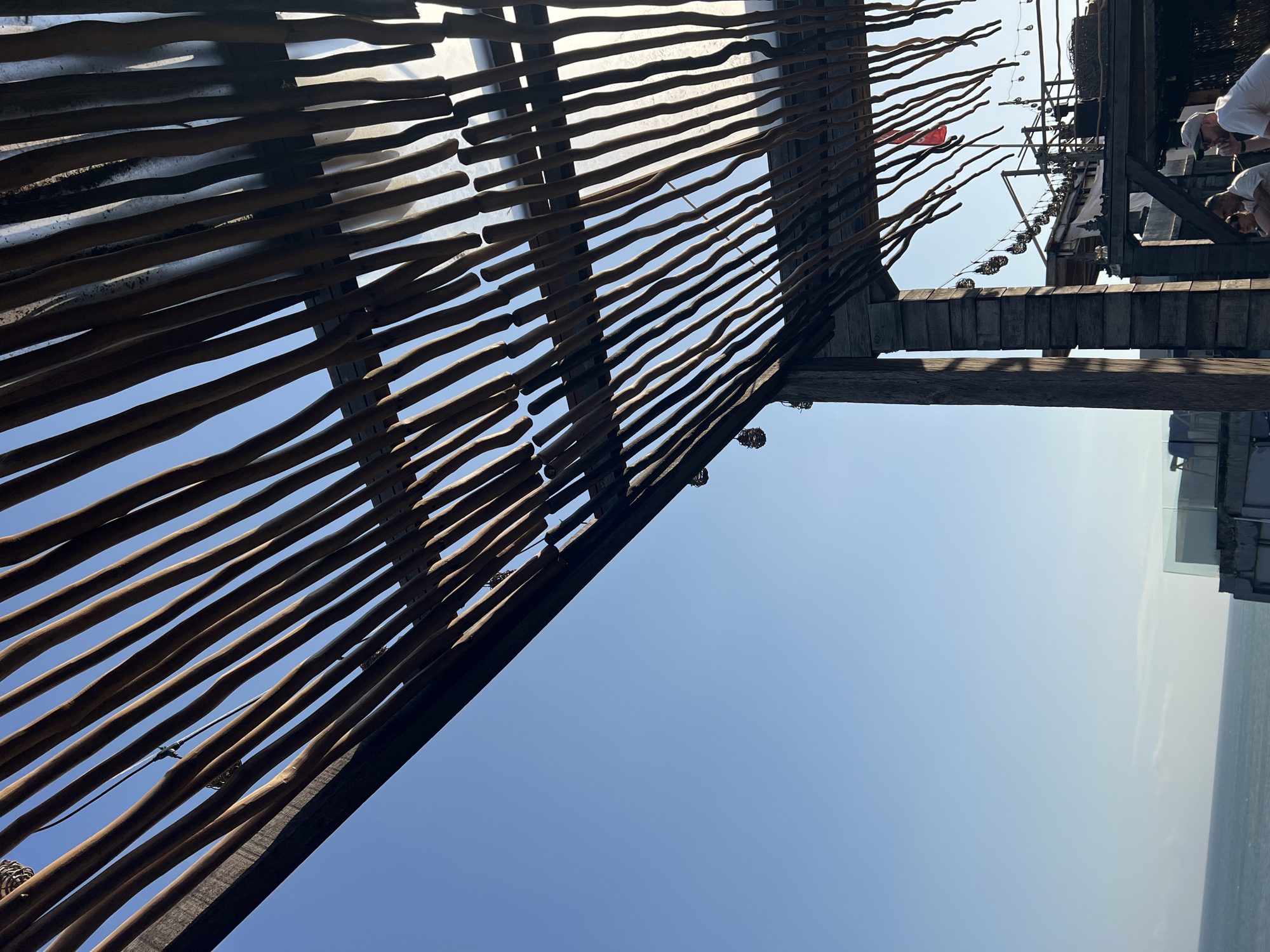 A low-angle shot captures an outdoor canopy structure with a slatted wooden roof, against a clear blue sky. The canopy is supported by rough-hewn wooden posts. Below the canopy, string lights with small, decorative shades are visible, along with glimpses of people. In the background, a red flag flutters, and the ocean stretches to the horizon with a building structure on the right. The scene suggests a rooftop or elevated dining or seating area in a coastal location, likely in Mengwi, Indonesia, given the context.