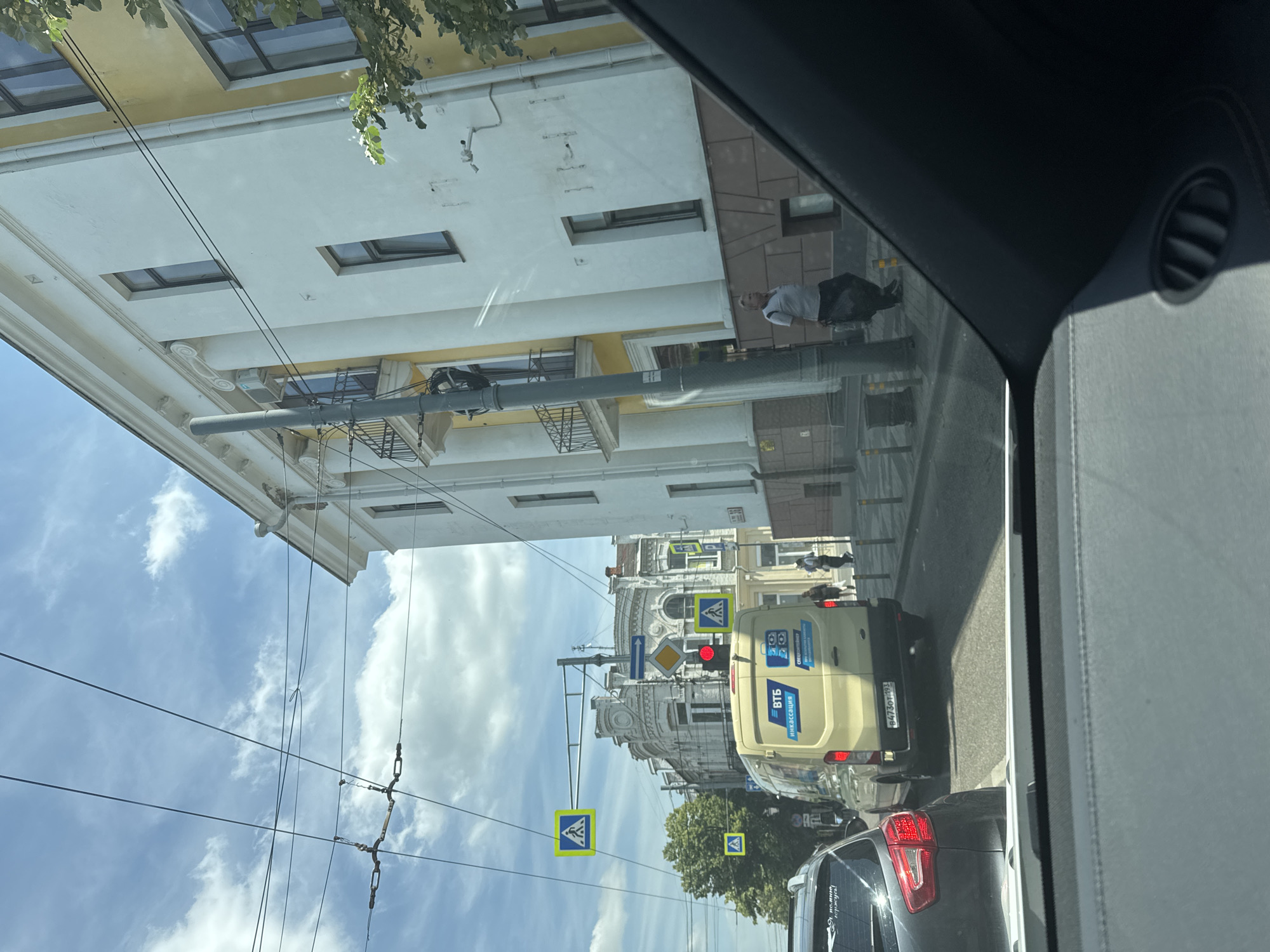 This is a photograph taken from inside a vehicle, looking out at a street scene in Krasnodar, Russia. The image is oriented vertically, and the contents appear upside down. It is daytime, with a bright blue sky and some clouds, suggesting fair weather.
The scene is a city street with several buildings visible on the left. The buildings are multi-story, with light-colored facades and some decorative architectural elements. Wires, likely for trams or power, crisscross the sky. On the right side of the image, the side of the vehicle from which the photo is taken is visible, including a dark interior with a black air vent.
In the middle ground, a yellow van is driving on the street. The van has blue text and logos on its side. One visible text reads "ВТБ" which is a Russian bank, and another text reads "ИНКАССАЦИЯ" meaning "cash collection". There is a license plate on the back of the van which reads "В475ОП 193". There are also several pedestrian crossing signs with a blue background and a white figure, indicating a crosswalk. A traffic light is visible, showing red. A person is walking on the sidewalk in the background.
In the foreground, the rear of a dark-colored car is visible. On its rear windshield, there is some text that is difficult to read clearly due to the image orientation and resolution.
The overall impression is a typical urban street view, possibly during rush hour, given the presence of multiple vehicles and pedestrian crossings.