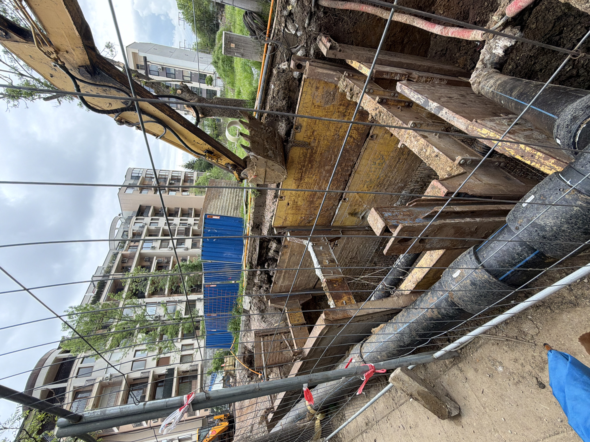 The image depicts a construction site in Sofia, Bulgaria, with a partially excavated trench. Yellow trench boxes are in place to support the trench walls. Large black pipes run along the side of the trench. An excavator with a yellow boom is visible. A temporary chain-link fence surrounds the site. In the background are residential buildings. The sky is overcast.