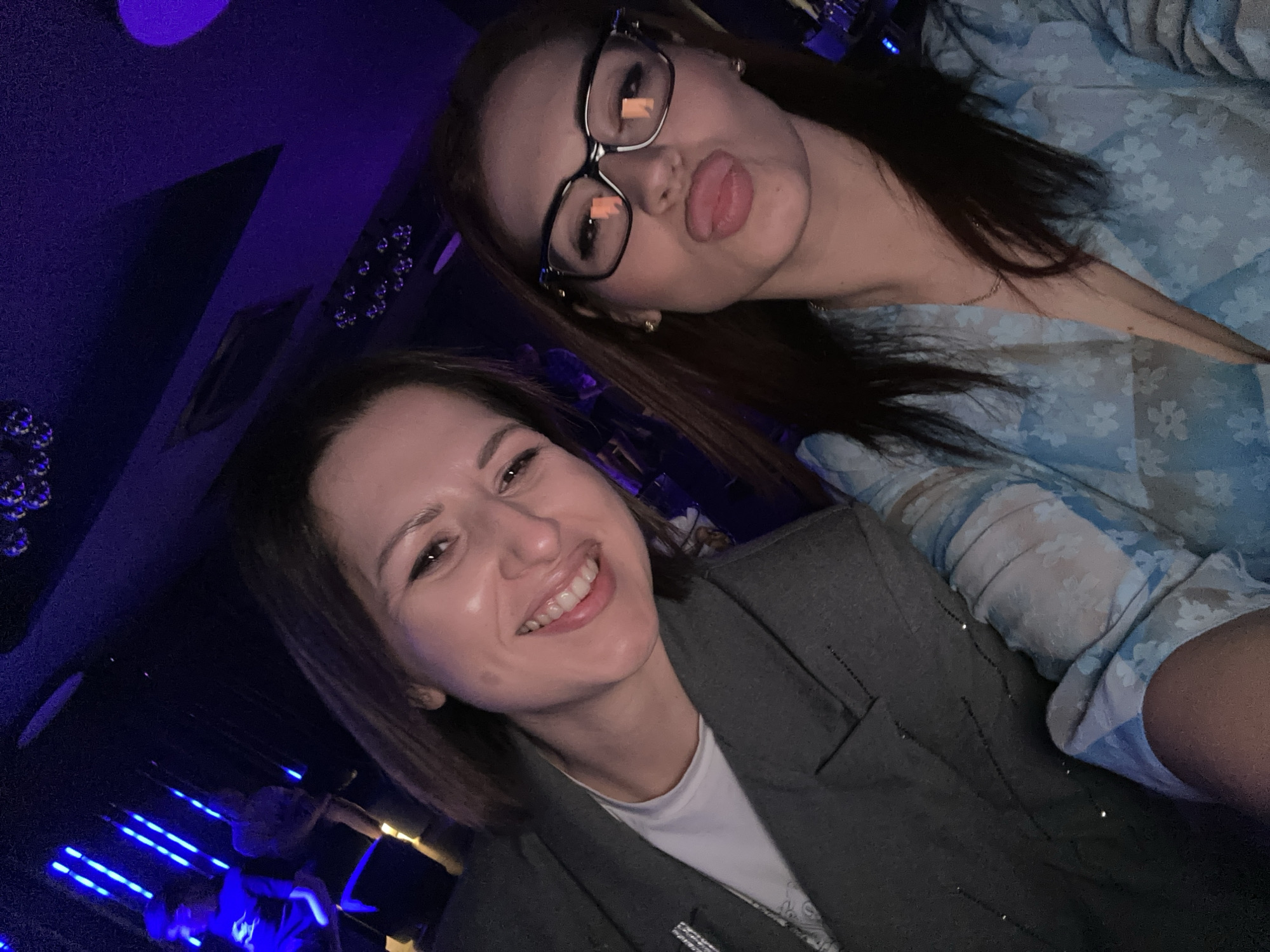 Two women are posing for a selfie indoors in Kazanlak, Bulgaria. The woman in the foreground has short brown hair and is wearing a grey blazer over a white shirt. She is smiling. The woman in the background has long brown hair, is wearing glasses, and is making a kissing face. She is wearing a light blue blouse with a floral pattern. The background features a blue neon light strip and dark furniture or decor.