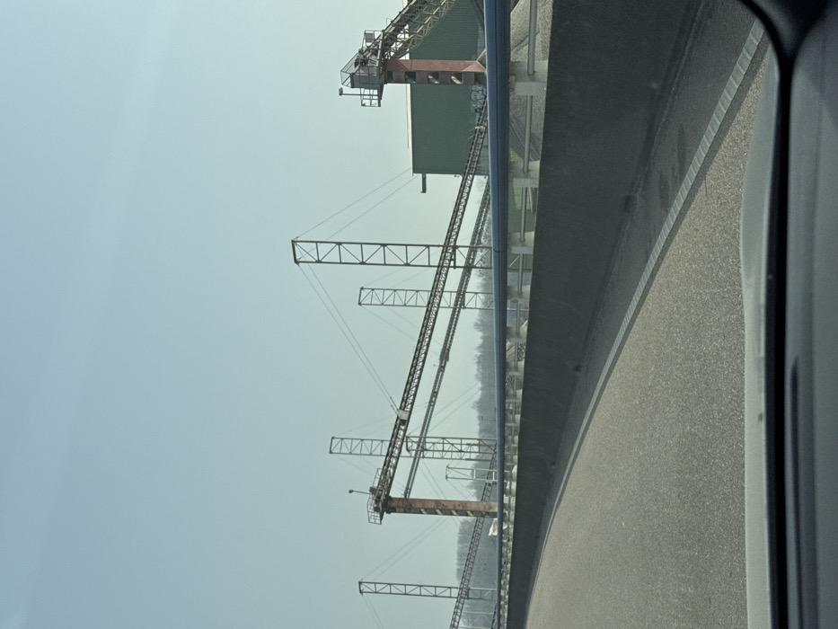 The image depicts an industrial material handling facility, located in Altenheim, Germany, observed from a vehicle's interior. In the foreground, the top edge of a vehicle dashboard is visible, along with a section of an asphalt road featuring a solid white edge line. A dark grey concrete barrier separates the road from an area containing piles of light-colored aggregate. Above this barrier, a series of light blue pipes are supported horizontally by white pedestals. The middle and background are dominated by an extensive network of elevated conveyor belt systems. These systems are supported by numerous tall, dark-colored lattice towers and substantial reddish-brown cylindrical structures. A large, dark green corrugated metal building is visible on the right, integrated into the conveyor infrastructure. Light-colored graffiti is present on a lower support section adjacent to the green building. A distant horizon line of leafless trees is visible beneath a clear, light blue sky. No individuals or active operations are discernible within the frame.