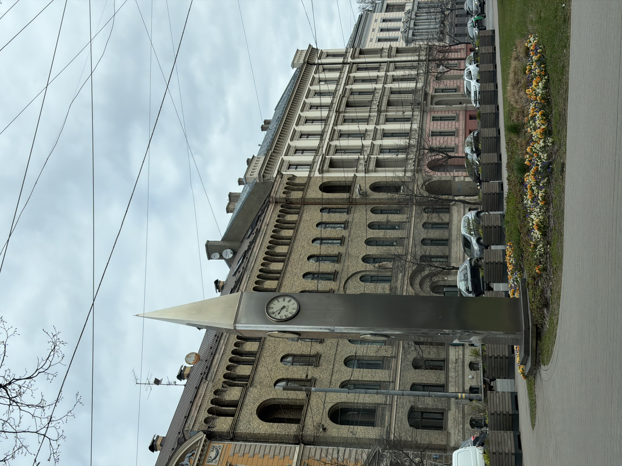 This image captures an urban street scene in Riga, Latvia, on an overcast day. A large, multi-story historical building dominates the left side of the frame, showcasing elaborate architecture. Its lower sections feature light-colored brick with numerous arched windows, while the upper floors are lighter in color with rectangular windows, topped by a detailed cornice.
In the mid-ground, a modern, metallic spire-like clock structure stands prominently. Its circular clock face displays Roman numerals and the word "RIGA," with the hands indicating approximately 12:45.
To the right, a paved sidewalk runs alongside a strip of green grass, which is brightly adorned with colorful flowers in shades of yellow, orange, purple, and white. Beyond the flowers and sidewalk, several cars are parked, including white, black, and green vehicles. Wooden benches are also visible along the pedestrian area.
The sky is uniformly grey and cloudy, with multiple overhead cables crisscrossing the upper left portion of the image. Bare tree branches suggest a season without full foliage, likely late winter or early spring. No people are visible, and the scene presents a quiet, typical view of a European city street.