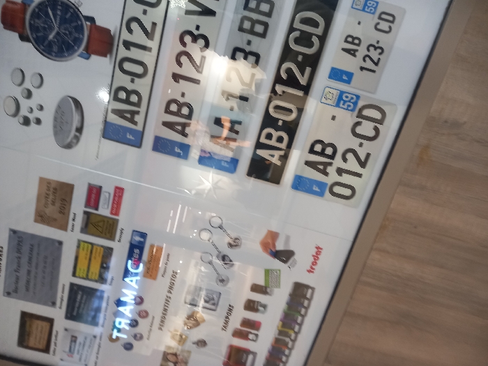 This image displays a brightly lit commercial display case or counter within a store located in Vendin-le-Vieil, France. The scene is dominated by a variety of customizable products and informational signage, presented on a white background with a light-toned wooden floor visible beneath.
On the upper left, multiple rectangular plaques are arranged. One prominently features "Docteur Franck DUPAS, MÉDECINE GÉNÉRALE," while another indicates "CUIVRE DES GUEULES 2019." Other plaques contain text and warning symbols, including a yellow triangle with an exclamation mark. Below these, a blue neon-style sign is visible, displaying "CAMART" (reflected as "TRAMAC"), with a smaller sign beneath reading "PENDENTIFS PHOTOS."
The lower left section showcases an array of small metal objects, including various keychains and pendants. Adjacent to these, a line of self-inking stamps in different colors is displayed, identifiable by the "trodat" brand. An interaction is depicted where a disembodied hand presses one of these stamps onto a white surface.
The upper right portion of the display features several circular metallic objects, resembling coins or tokens, of varying sizes. A wristwatch with a blue face and a brown leather strap is also prominently displayed.
The lower right section presents multiple sample French vehicle license plates. Examples include white plates with black lettering such as "F AB-012-CD," "F AB-123-V," and "F AA-123-BB," many featuring the European 'F' insignia and the regional department number '59'. A black license plate with white lettering, displaying "AB-012-CD," is also present.
The entire display surface exhibits reflections, including fragmented images of the surroundings and possibly the person capturing the image.
