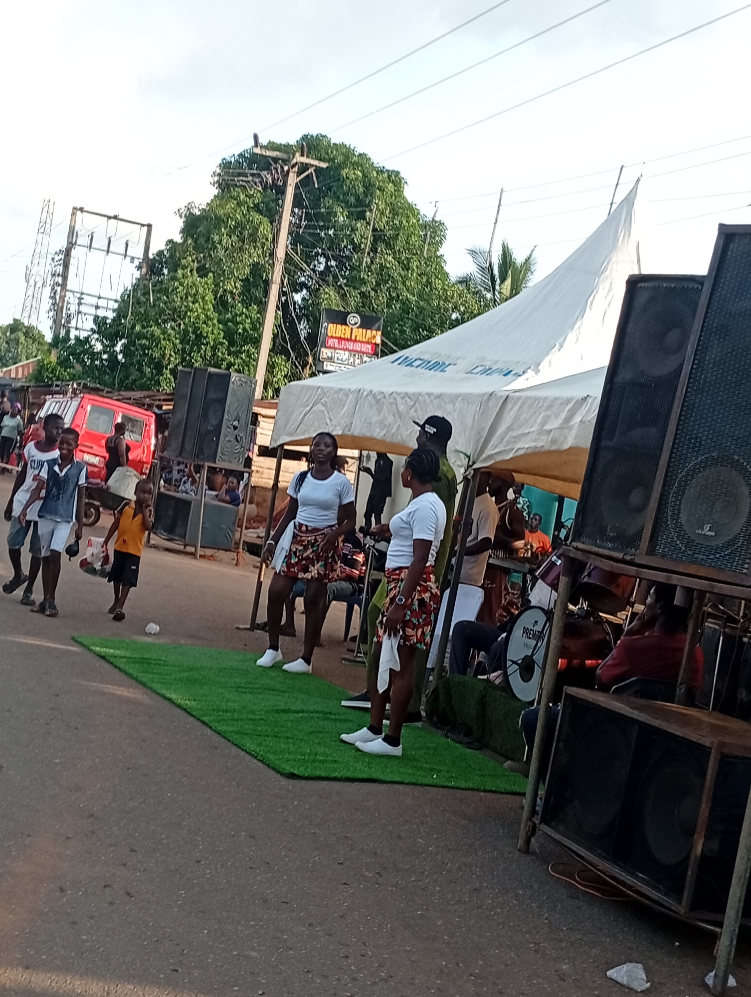 The image depicts an outdoor scene, possibly in Nsugbe, Nigeria, featuring a street event with music and a crowd. In the foreground, a bright green, artificial turf mat is laid on the asphalt road, with several people standing and walking on it. To the right, large speaker cabinets are set up, some on a metal frame, indicating a sound system for the event. A white tent or canopy stretches across the middle ground, providing shade. Underneath the tent, a band appears to be performing, with drums and other musical instruments visible. Several individuals are seen in the background, some interacting with each other and others engaged in the festivities. A red van is parked on the left side of the road, and electrical poles and wires are present in the distance. The overall atmosphere suggests a lively community gathering or celebration.