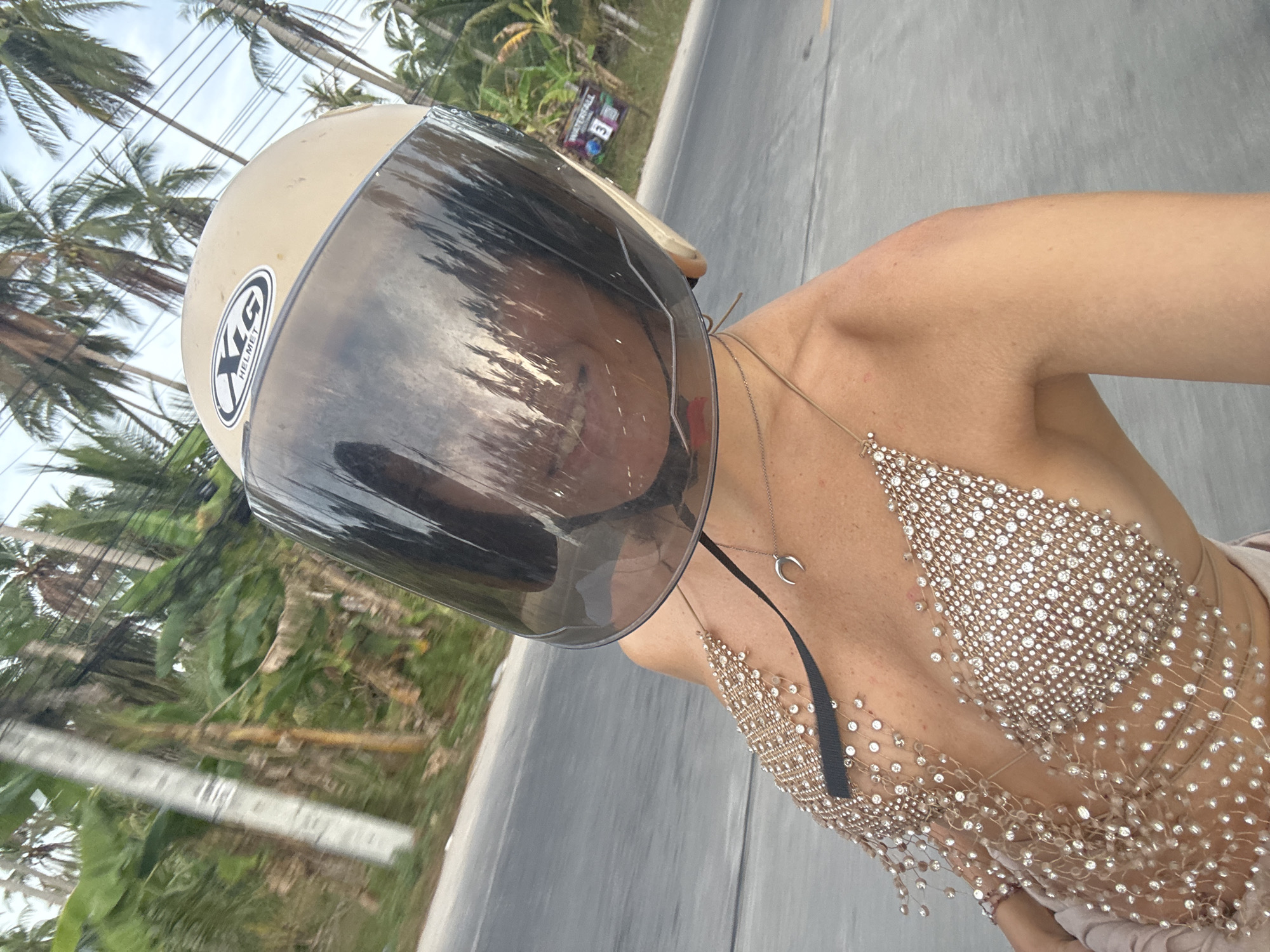 A woman is on a road in Moo 4, Thailand, wearing a beige helmet with a tinted visor, a sparkling beaded top, and a necklace. The background shows palm trees and a roadway. In the distance, a sign saying "WATERFALL" can be seen next to the roadway.