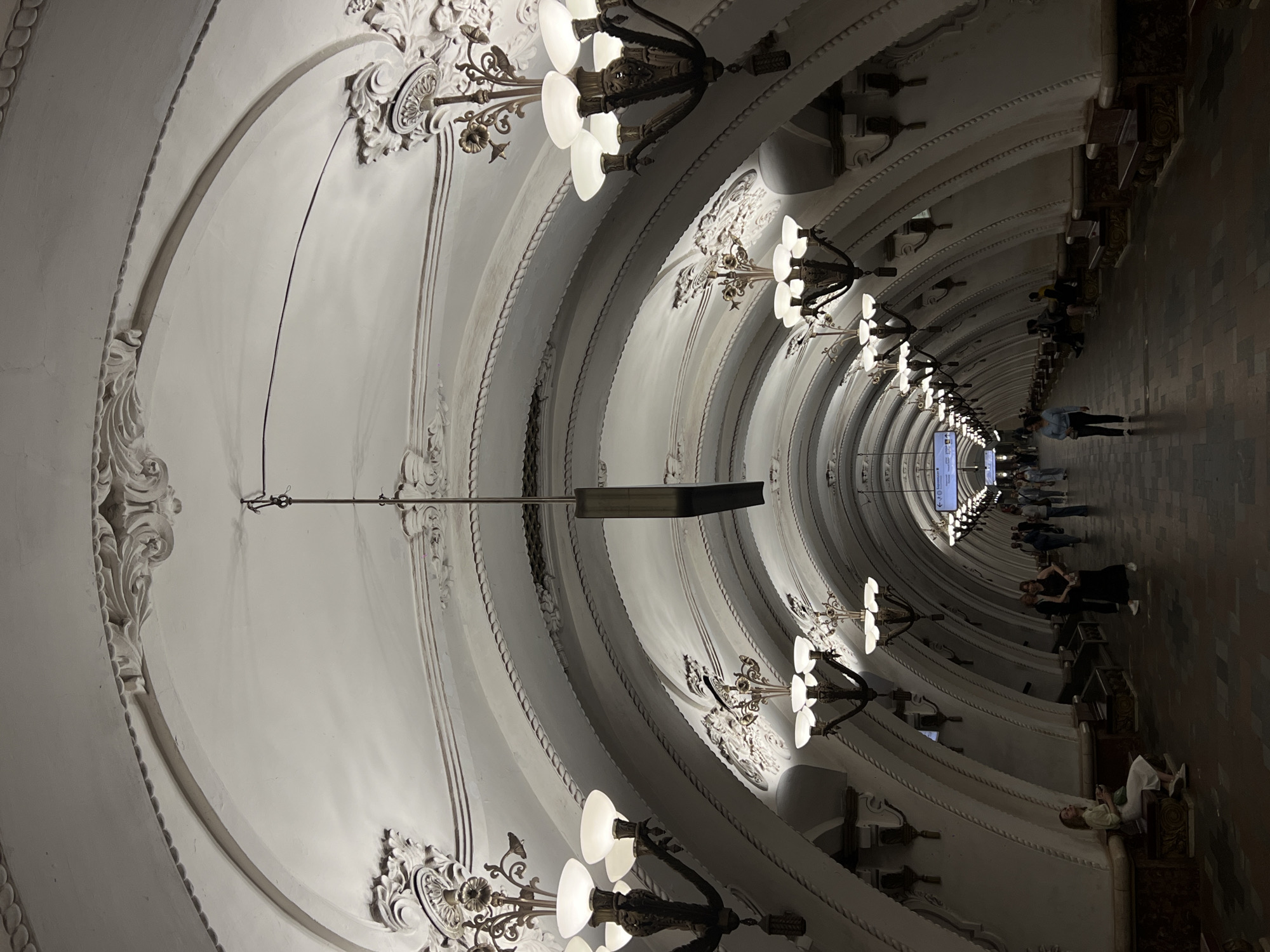 This image shows the interior of a Moscow metro station. The architecture features a series of archways with ornate, white-painted moldings and decorative chandeliers. Passengers are visible walking and sitting along the platform. The overall ambiance is dimly lit, with the light focused on the archways and chandeliers.