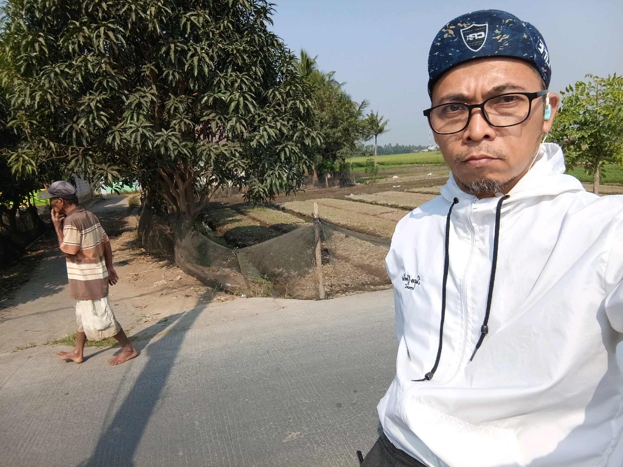 In Rawa Boni, Indonesia, a man wearing glasses, a blue cap, and a white hooded jacket is taking a selfie. He has light blue earbuds in his ears and a goatee. In the background, another man walks barefoot on a paved path. He wears a striped shirt and shorts. The environment consists of a large tree, cultivated fields, and lush vegetation under a clear sky.