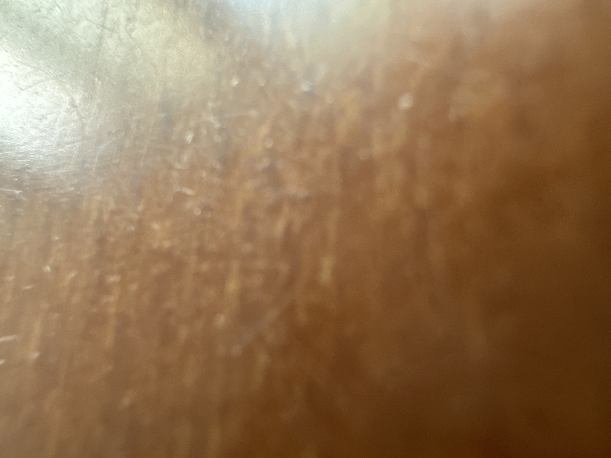 The image depicts a close-up, out-of-focus view of a light brown, textured surface, possibly wood grain, with subtle variations in color and small, lighter specks that could be dust or imperfections. The lighting creates a bright, reflective sheen across a portion of the surface, particularly in the upper right quadrant. The overall impression is of a generic, indoor surface, devoid of any discernible landmarks or context that would allow for identification of its location within Belgrade, Serbia, or any other specific place. There are no people, objects, actions, or interactions visible.