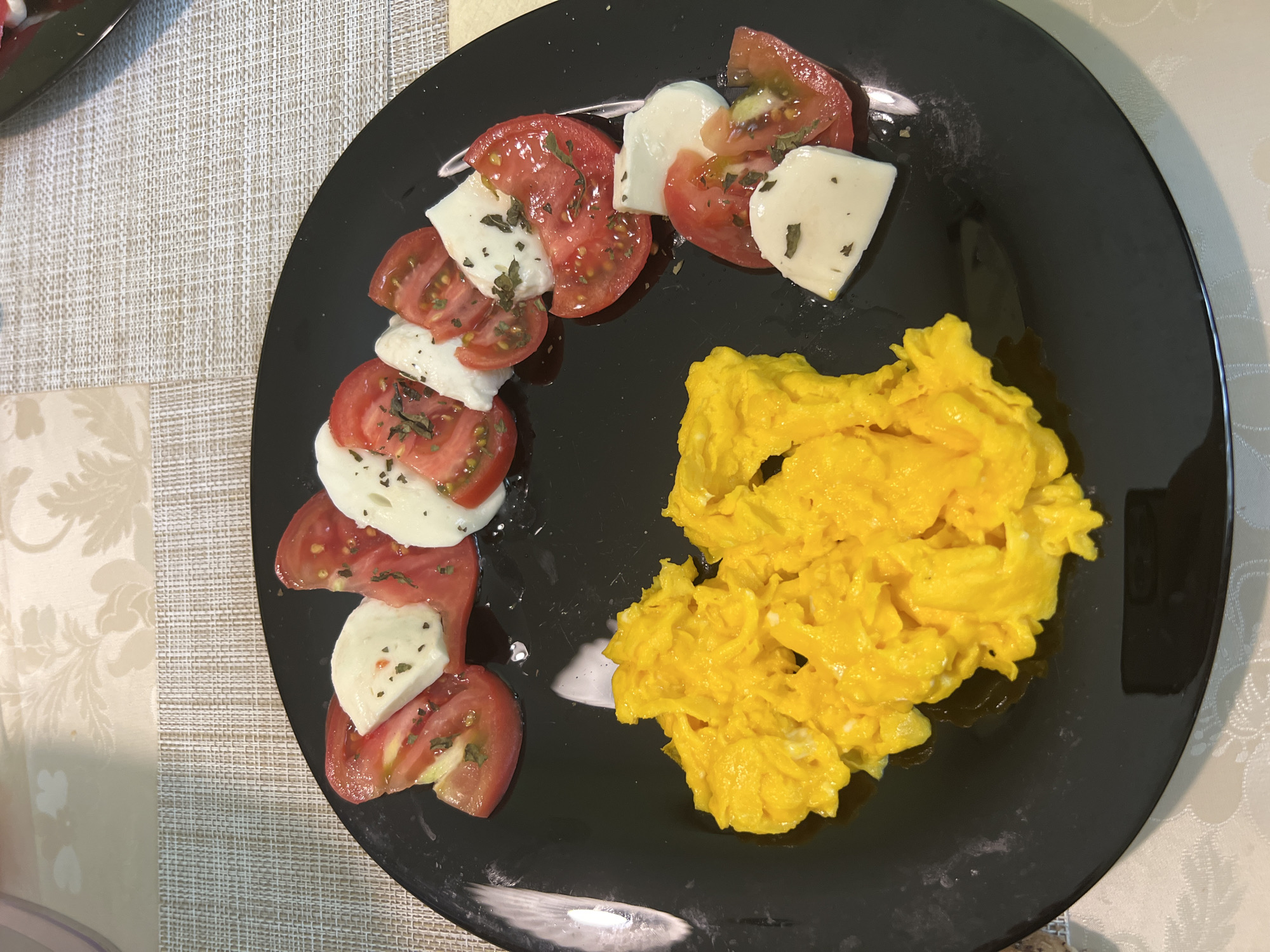 A close-up, high-angle shot captures a freshly prepared meal served on a glossy black circular plate, presented on a light beige woven placemat atop a subtly patterned tablecloth in the city of Vratsa, Bulgaria. The plate features two distinct components: a vibrant yellow mound of scrambled eggs occupying the left half, and a linear arrangement of sliced red tomatoes interspersed with thick cuts of white mozzarella cheese extending across the right side. Both the tomatoes and mozzarella are generously seasoned with dried green herbs, likely basil or oregano, and appear to be drizzled with a clear oil or dressing. No individuals are present, and the scene is illuminated by overhead artificial lighting, reflecting faintly on the plate's surface.