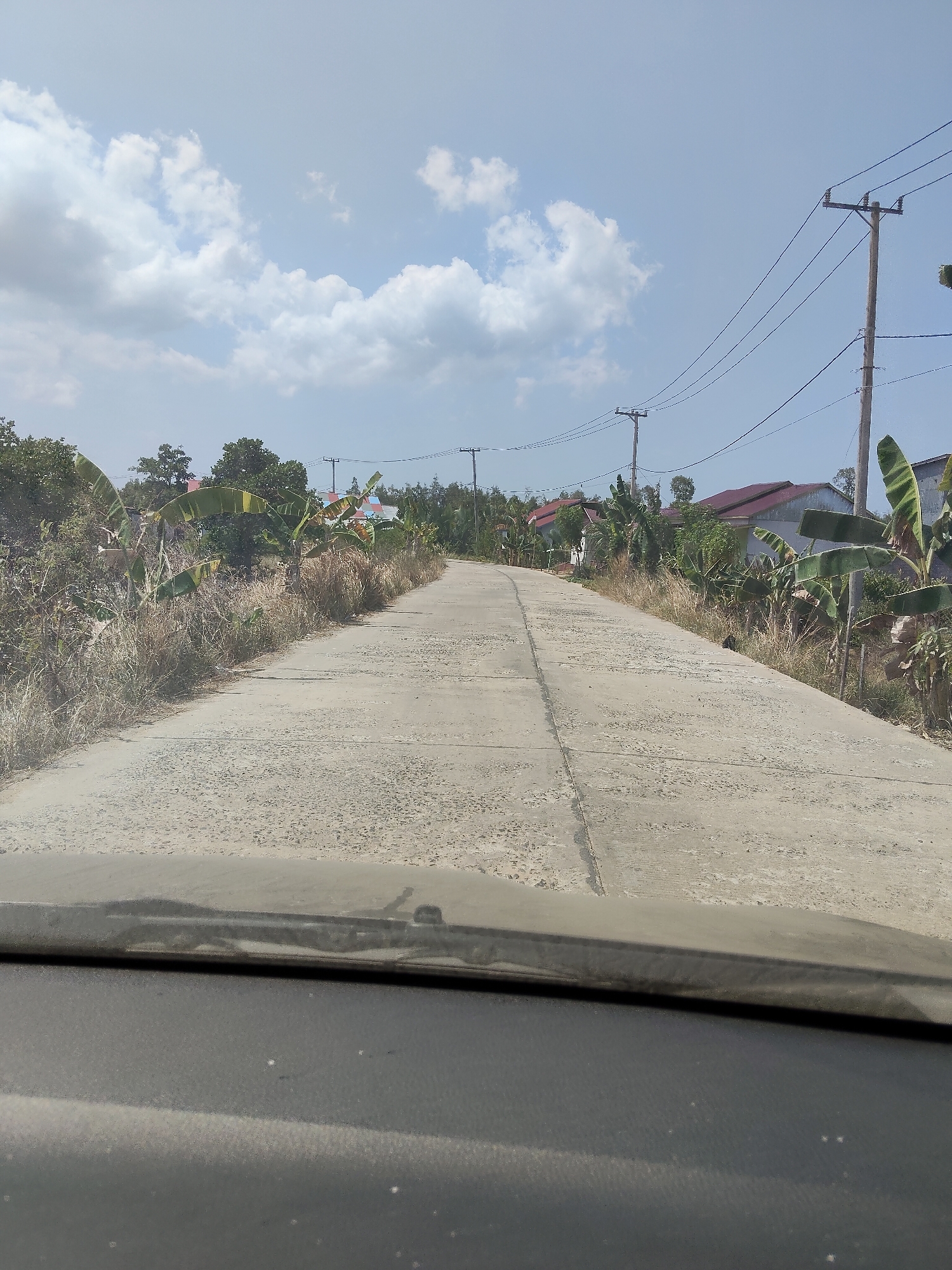 The image is a forward-facing view from a vehicle, looking down a paved, two-lane road. The road is in fair condition, with visible cracks and some unevenness. On both sides of the road, there is dry, tall grass and scattered vegetation, including banana trees. Beyond the vegetation, several houses with red roofs are visible, along with utility poles and overhead power lines. The sky is blue with scattered white clouds. The scene appears to be in a rural or semi-rural area of Lori, Indonesia, likely a residential street. There are no people visible in the image. The lower portion of the image shows the vehicle's dashboard and windshield.