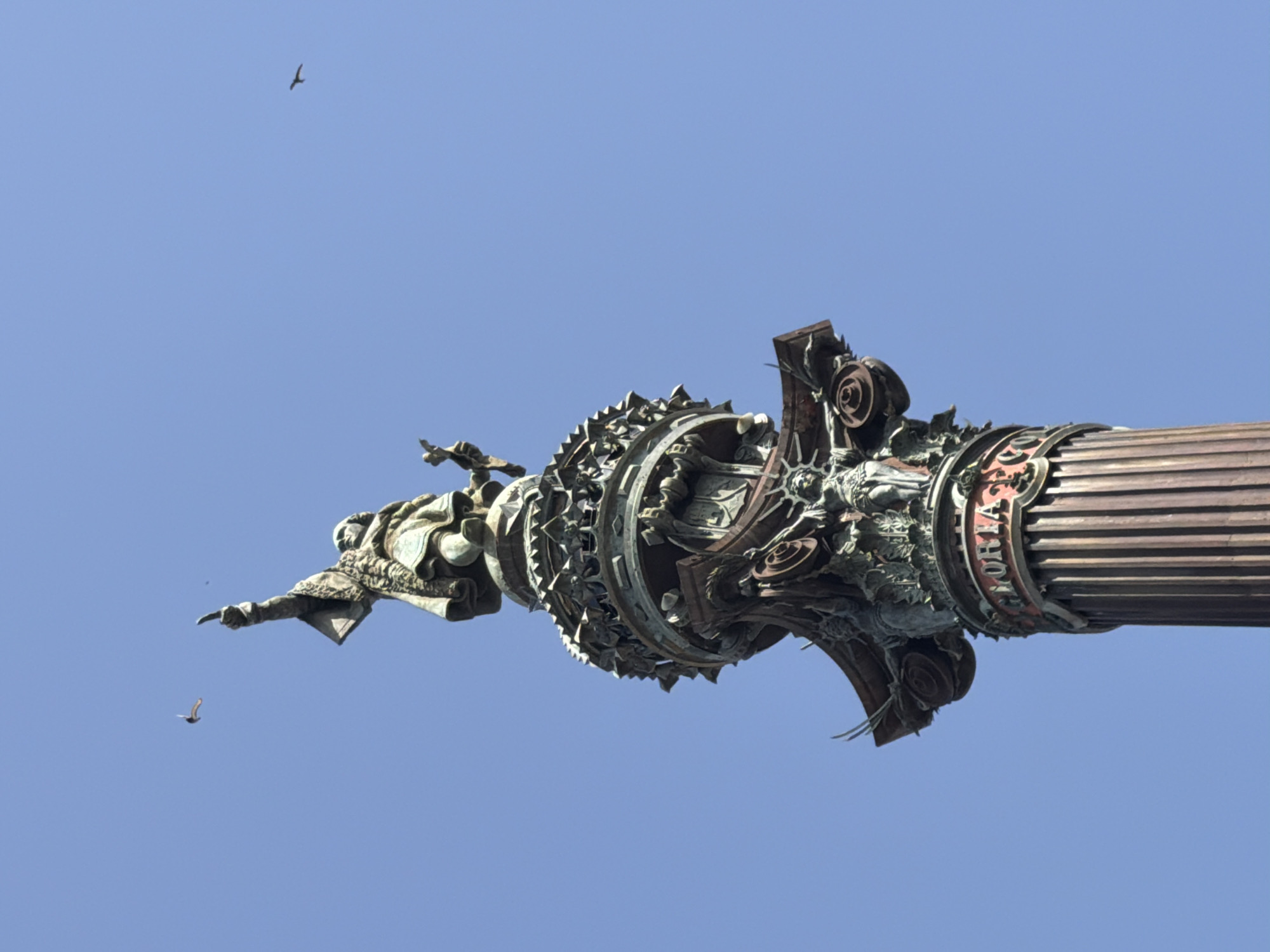 The image shows the Monument to Columbus in Barcelona, Spain, a towering bronze statue located at the end of Las Ramblas at Port Vell. The statue of Christopher Columbus stands atop a fluted column, pointing outwards. The monument features intricate sculptural details, including figures and decorative elements around the column's capital. In the bright blue sky above the monument, two birds are visible in flight. The lower portion of the column has the word "GLORIA" inscribed on it in red lettering. The photograph is taken from a low angle, looking up at the monument against the clear sky.