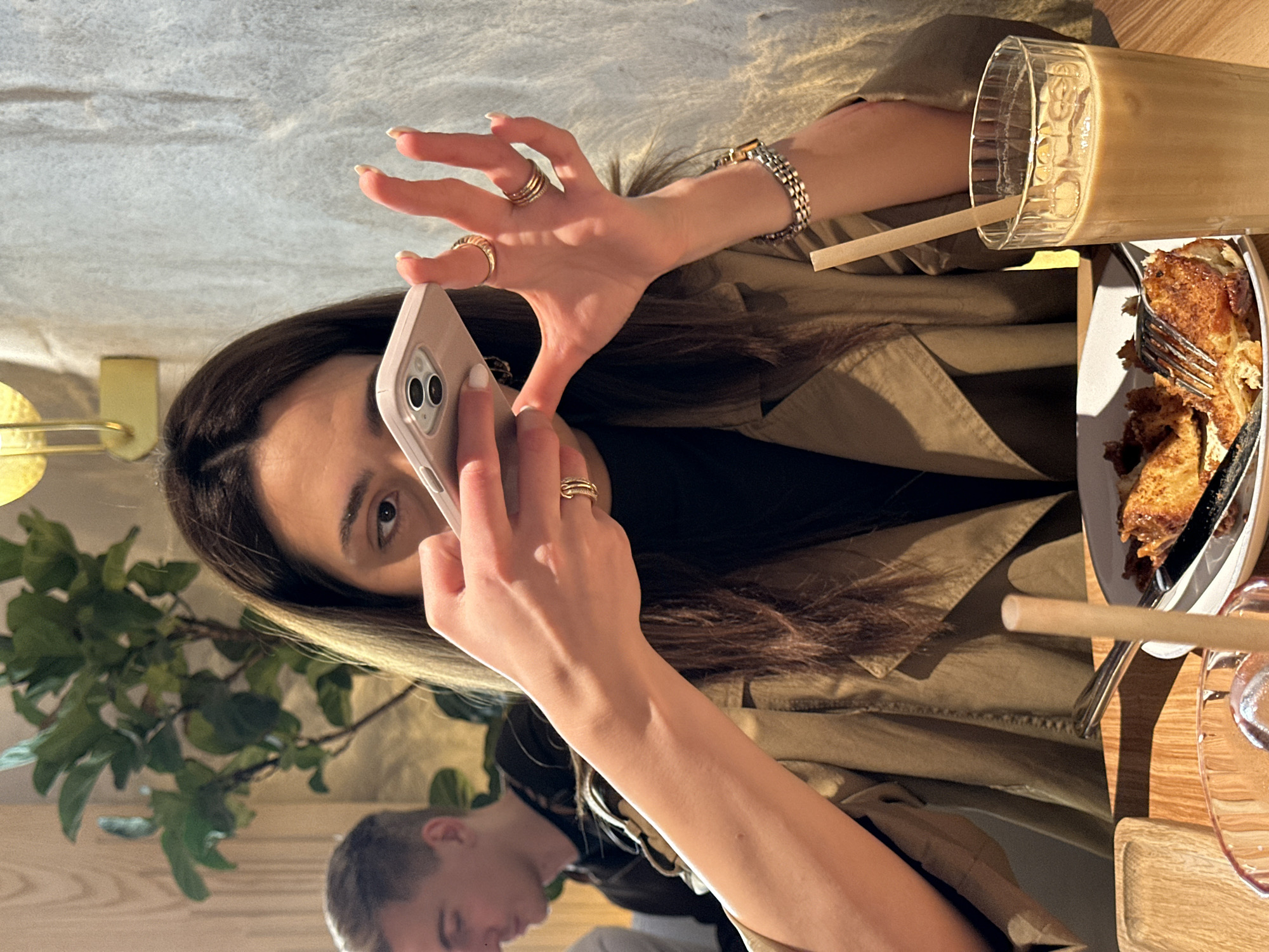 A woman with long brown hair is holding a light pink cell phone and taking a picture. She has multiple rings on her fingers and is wearing a gold bracelet. She is wearing a tan trench coat. In front of her is a glass of iced coffee with a straw and a plate of food with a fork. There is a man with short hair in the lower left corner of the frame. A green leafy plant is visible behind the woman. The scene is likely inside a cafe or restaurant. The location is identified as Yerevan, Armenia, based on external knowledge.