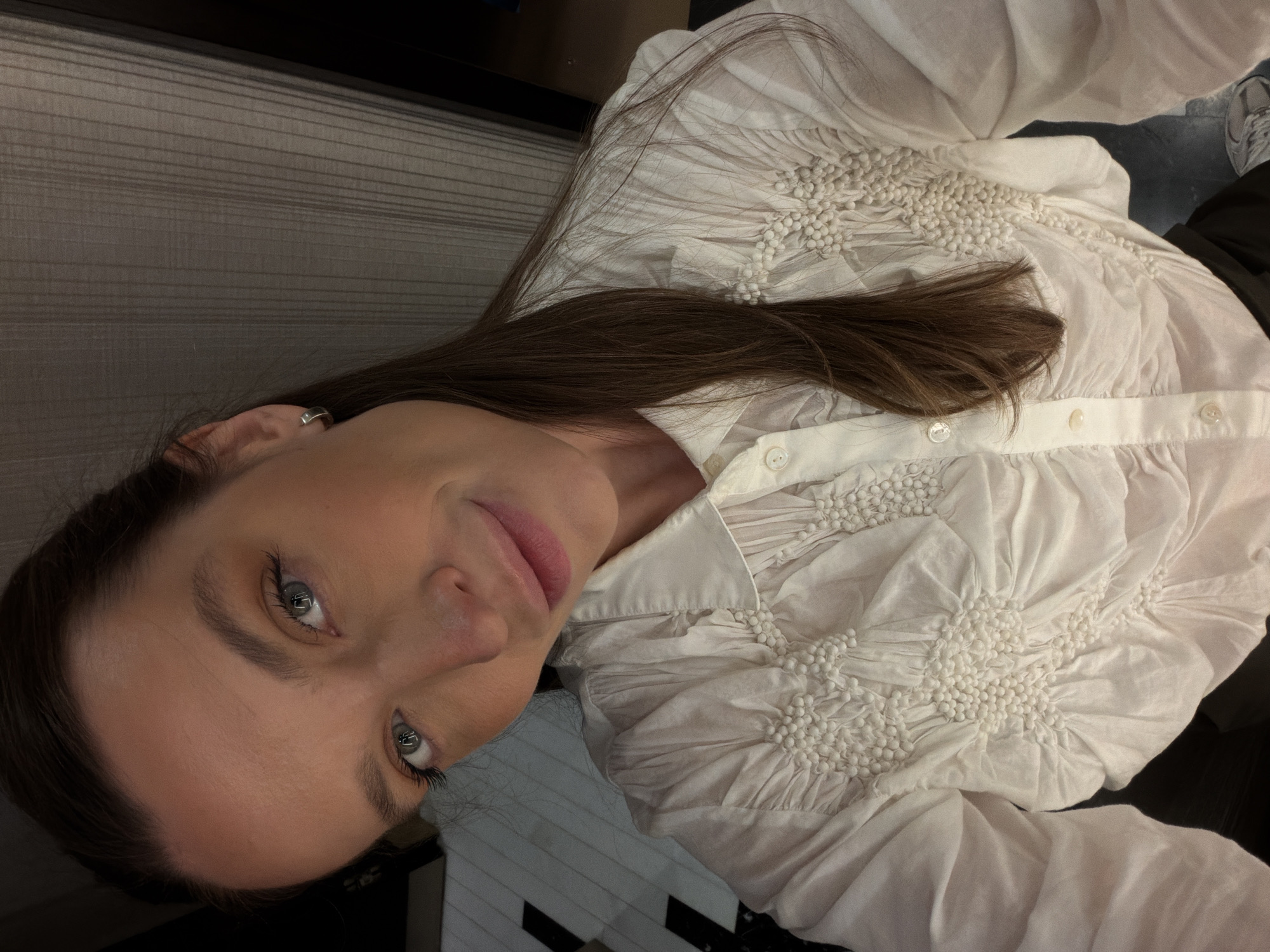 A woman with brown hair and blue eyes is captured in a selfie-style shot. She is wearing a white button-down shirt with decorative detailing. The background shows a hint of interior with what appears to be blinds, suggesting the photograph was taken indoors. While the image does not provide sufficient information to confirm the location in the city of Budva, Montenegro, it can be stated that the setting is indoors.