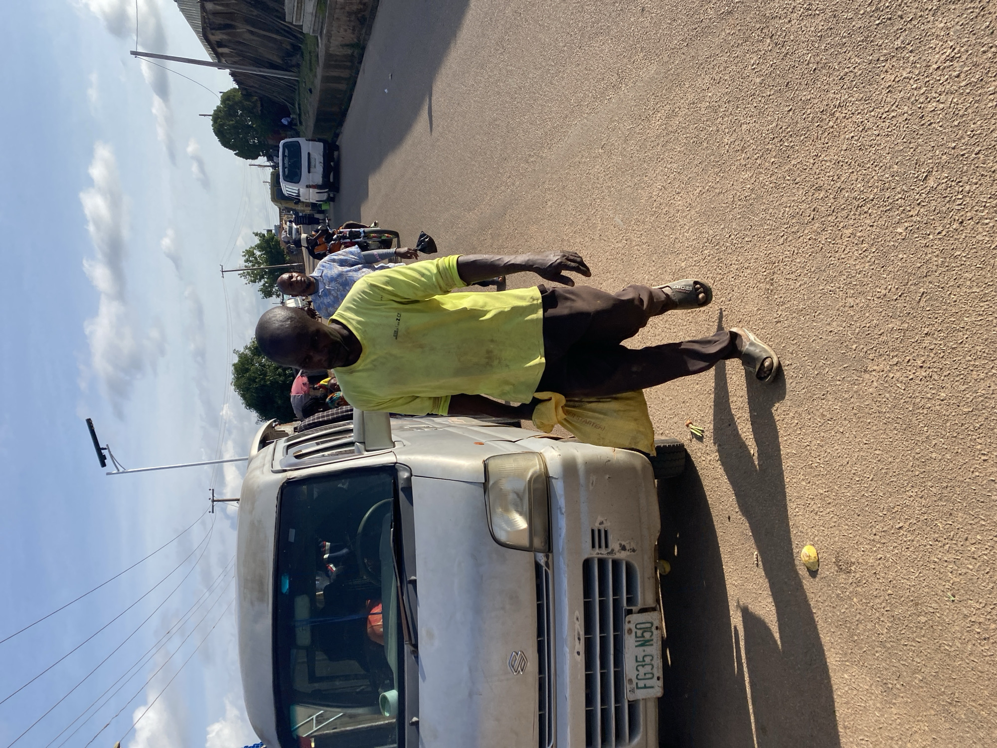 A man in a fluorescent yellow long-sleeved shirt and dark pants walks down a street in Gura Top, Nigeria, carrying a yellow plastic bag. He is walking in front of a silver Suzuki van with a license plate that reads "FG35 N50". The street is paved with a reddish-brown surface, and there are several other people and vehicles in the background, including a motorcycle and another van. The sky is blue with scattered white clouds. The scene appears to be a busy street with signs of commercial activity.