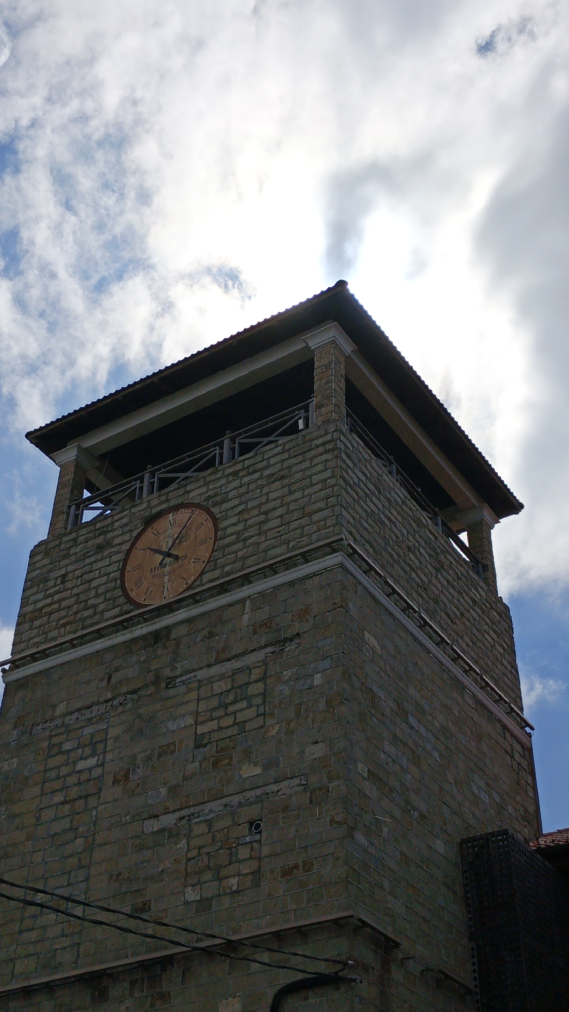The image shows a low-angle view of the clock tower in Abrau-Dyurso, Russia. The tower is constructed of light brown stone blocks and features a large clock face. The hands of the clock show approximately 12:45. The tower has an overhanging roof with an open viewing platform below. The sky is visible behind the tower, with clouds and some blue sky. A few power lines are visible in the bottom left of the frame.