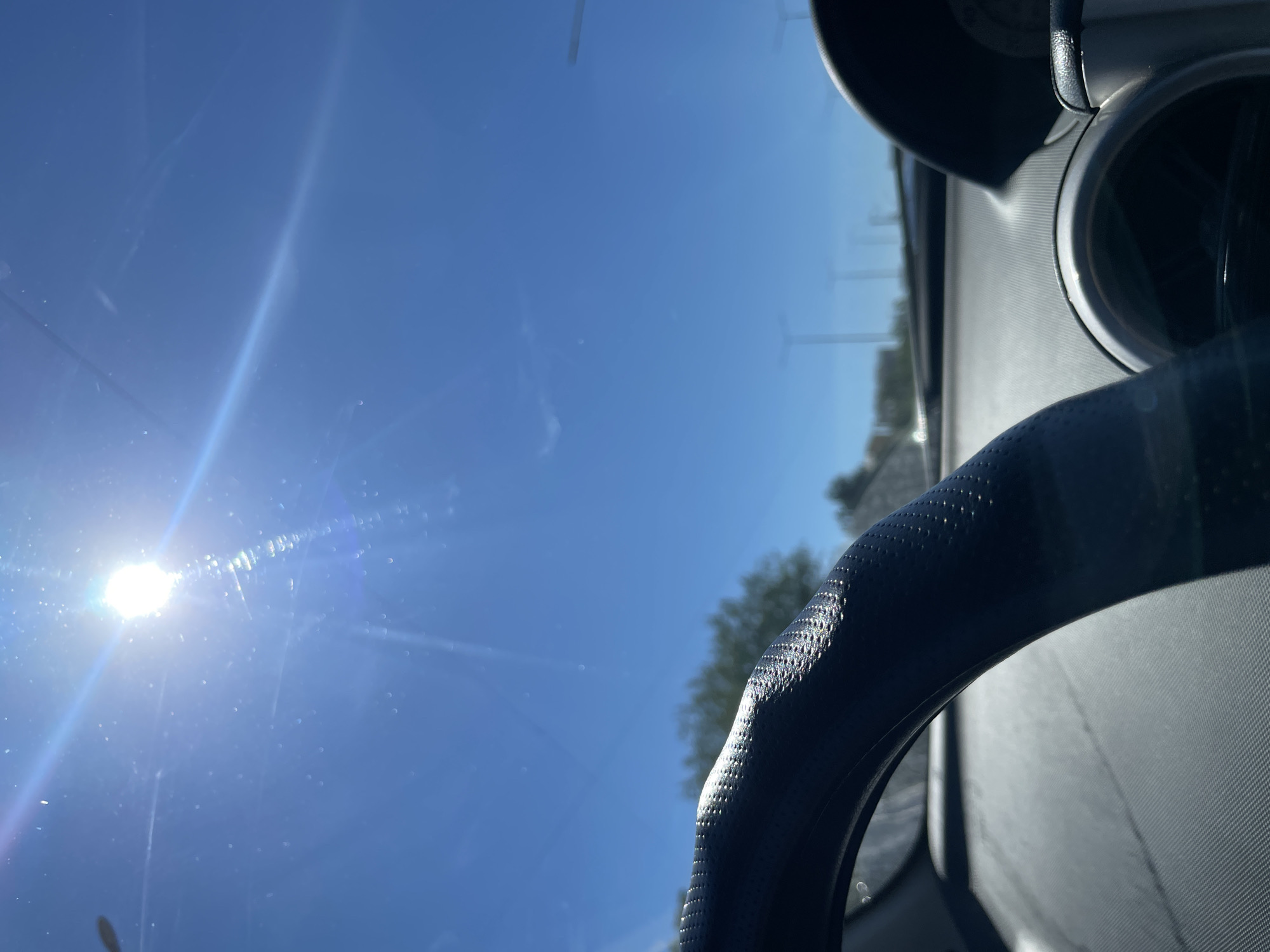 The image shows a view from inside a car, looking out through the windshield on a bright, sunny day. The sun is high in the sky, casting strong glare and lens flares across the scene. The foreground is dominated by a portion of a black steering wheel, textured with a pattern of small dots. To the right, parts of the car's dashboard are visible, including a dark curved surface and the edge of a circular gauge.
Beyond the car's interior, the view through the windshield reveals a clear blue sky. In the distance, trees with green foliage are visible, and a highway or road with lampposts can be seen. The overall impression is that of a car driving on a clear day, likely on a highway or a road with sparse urban development.
The location is specified as Aktau, Kazakhstan. While the image itself does not contain specific visual cues to pinpoint the exact location within Aktau, the context suggests it is a road in or near the city. The presence of the highway and lampposts is consistent with urban or suburban road infrastructure. The sunny weather indicates a typically clear day in the region. There are no people visible in the image, and the focus is on the car's interior and the external environment.