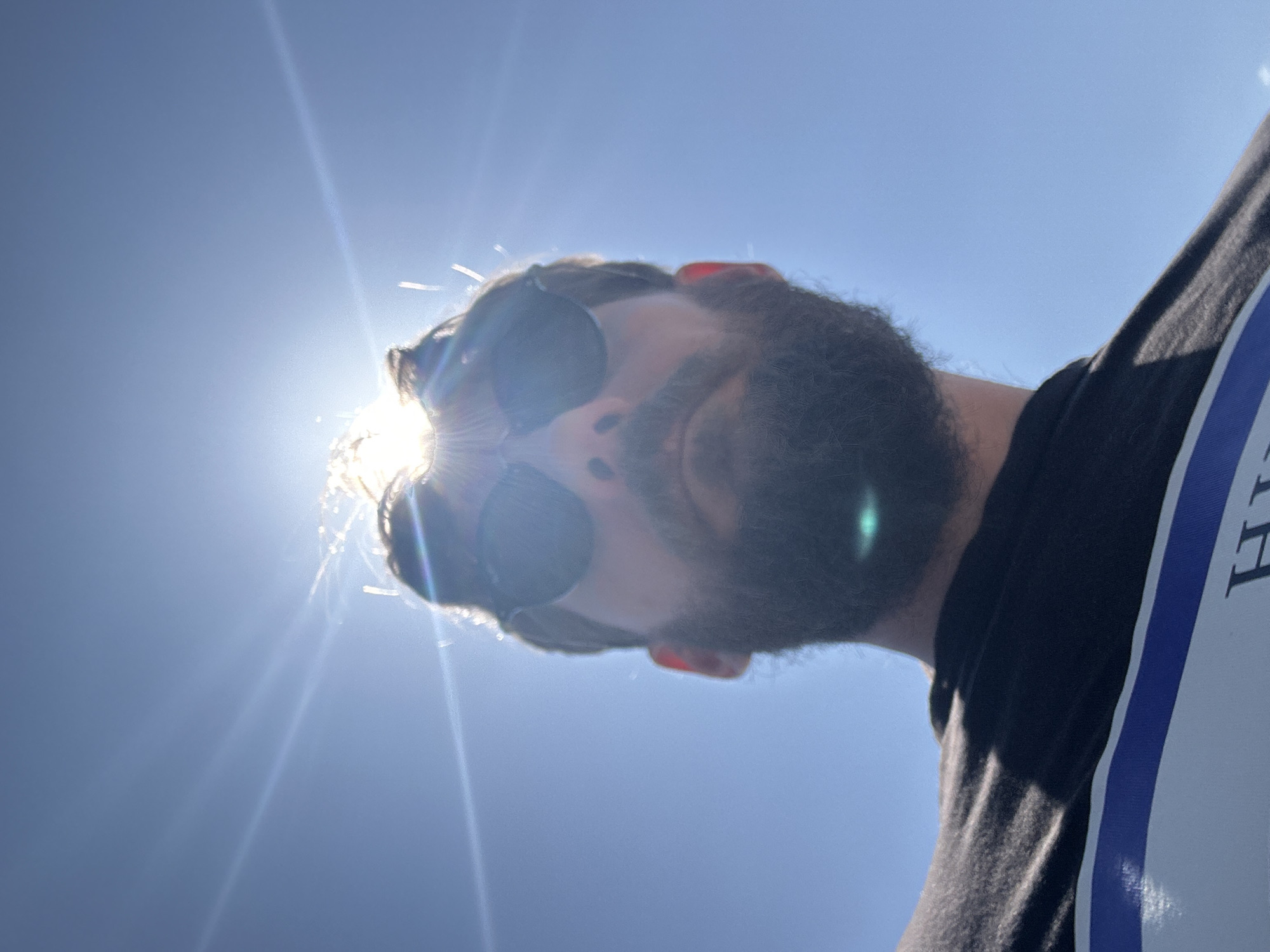 The image is a low-angle portrait of a person with a beard and sunglasses, captured against a clear, vibrant blue sky. In its original orientation, the person's head is on the right side of the frame, looking towards the bottom-center. The sun is positioned behind and slightly to the left of the person's head, creating intense backlighting, prominent lens flares, and a bright halo effect around their hair and upper head.
The subject is a male with a dark, full beard and dark hair. They wear round, dark-lensed sunglasses, which obscure their eyes and much of their face due to the harsh backlighting. They are wearing a black t-shirt that features a vertical blue and white stripe along the right side of the chest. A partial letter, likely "H," is visible on the white stripe.
The sky is entirely cloudless, indicating a bright and sunny day, possibly mid-day or afternoon given the sun's high intensity and position. The overall impression is one of clear weather and outdoor conditions. While specific landmarks are absent, the bright, clear sky is characteristic of sunny coastal regions like Fethiye, Turkey. The perspective suggests the photo was taken from below, looking up at the person and the sky.