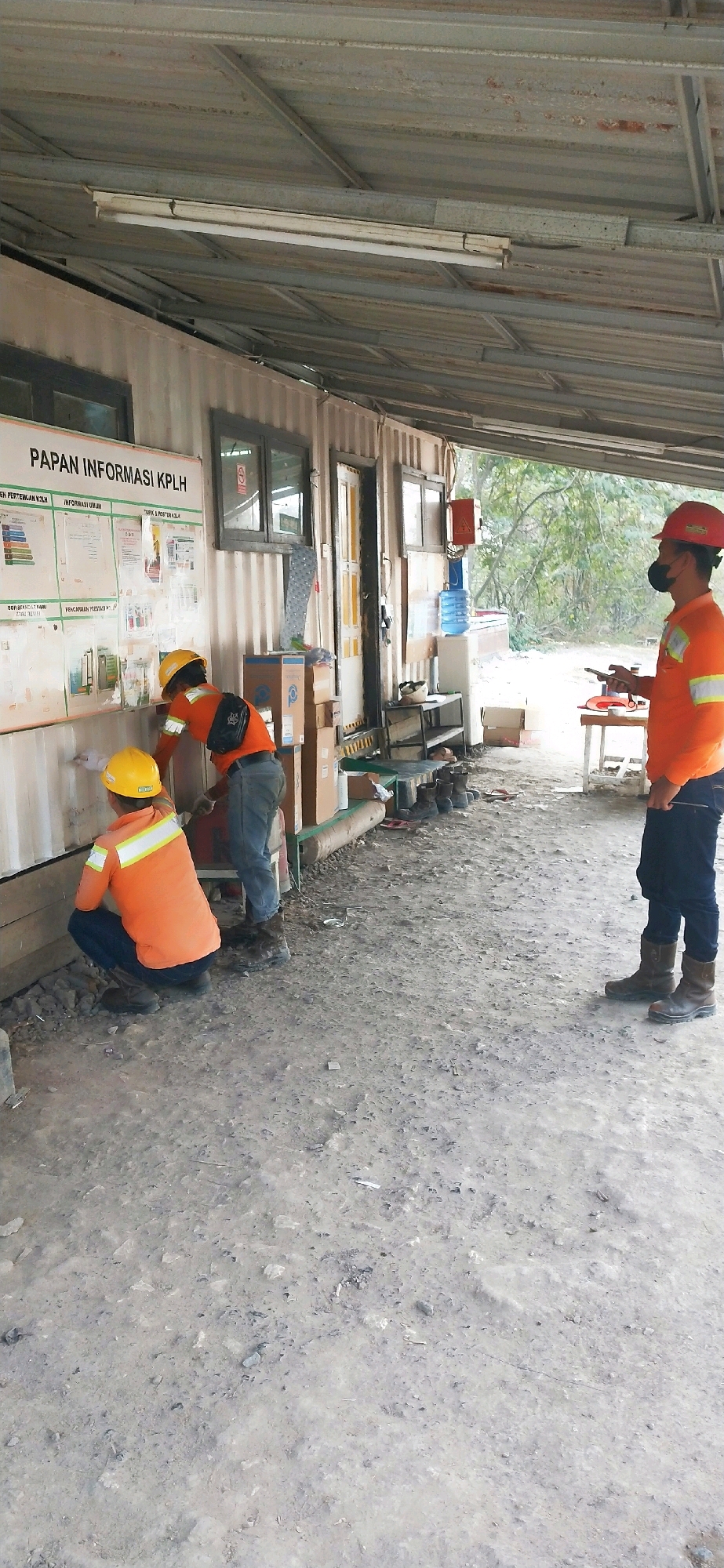 The image depicts an outdoor scene in Paringin, Indonesia, likely a construction or work site, characterized by a corrugated metal structure serving as a temporary building or shelter. In the foreground, the ground is covered with gravel and dirt, with some scattered debris.
There are three individuals present, all wearing orange high-visibility work shirts and hard hats, indicating they are likely construction workers or site personnel. Two of the individuals are positioned in front of the corrugated metal structure, engaged in what appears to be maintenance or cleaning work on the exterior wall. One of them is crouching down, while the other stands slightly behind, both holding tools or brushes.
The third individual is standing to the right of the frame, facing away from the structure and towards the viewer's left. This person is also wearing an orange work shirt and a red hard hat and appears to be holding a mobile phone and possibly another object in their hands, suggesting they might be observing, directing, or documenting the work.
The corrugated metal structure itself has windows and doors, and a large information board labeled "PAPAN INFORMASI KPLH" is mounted on the exterior wall, displaying various charts and documents. Various items, including boxes, a water dispenser, and other equipment, are visible around the structure, further suggesting a working environment. The overall atmosphere is industrial and functional.