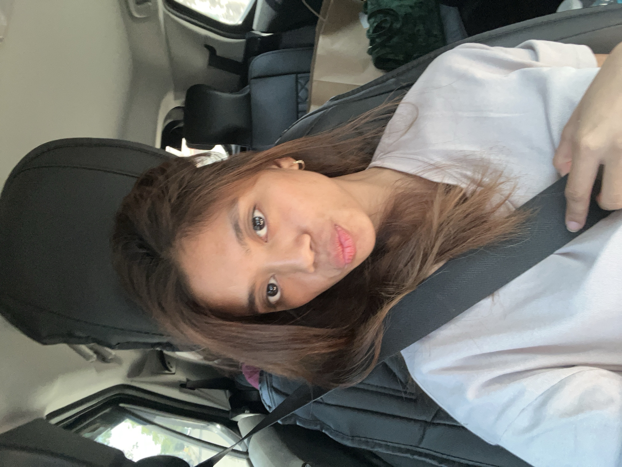 A female individual is depicted in a self-portrait taken inside a vehicle. She has long, dark brown hair and is looking directly at the camera with her lips puckered. She is wearing a light-colored, short-sleeved top and a black seatbelt across her chest and shoulder. A small gold-colored hoop earring is visible on her right ear. Her right hand is positioned on the seatbelt. The car's interior features dark grey or black upholstered seats and headrests. A light-colored ceiling is visible above. In the background, a paper bag and a dark green, textured item are present on the rear passenger seat. Through the vehicle's windows, a blurred exterior environment, suggestive of daylight and foliage, is visible. The scene is located in Davao City, Philippines.