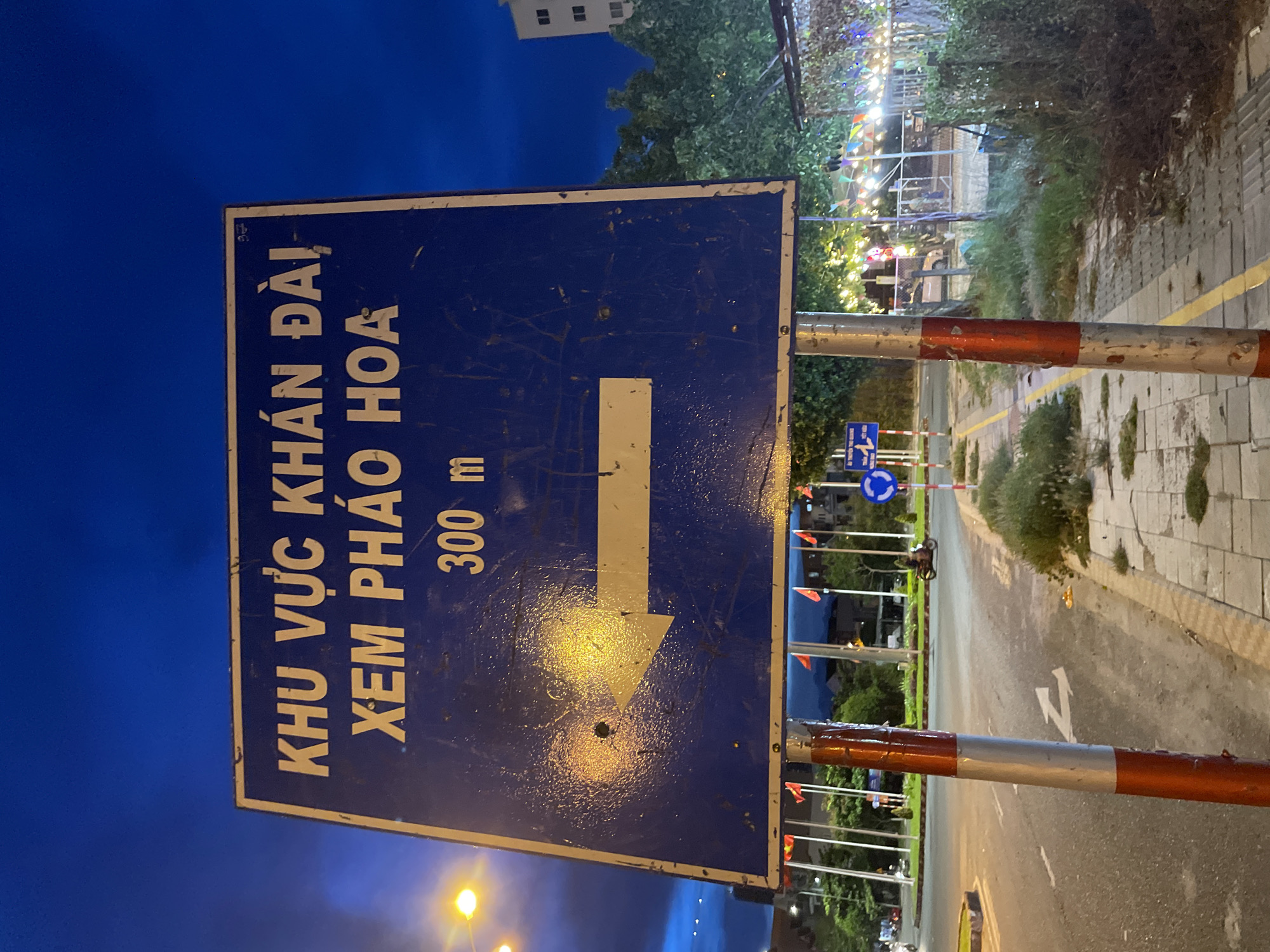 A blue directional sign with white text and a white arrow is mounted on two red and white striped poles. The sign reads "KHU VỰC KHÁN ĐÀI XEM PHÁO HOA" and "300 m" with an arrow pointing left. The background shows a street at dusk with streetlights and flags. In the distance, there are trees and buildings. A motorcycle is visible on the road. The scene appears to be in Đà Nẵng, Vietnam, likely near a location for viewing fireworks.