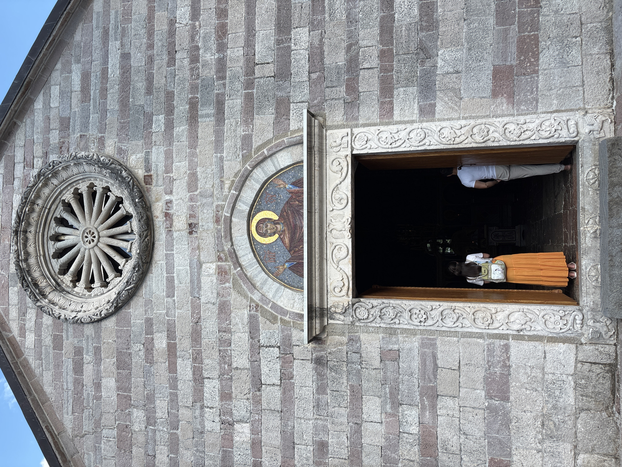 A vertical, eye-level outdoor shot shows the stone facade of a building, likely a church, in Budva, Montenegro. The wall is constructed from rectangular, grey and pinkish-grey stones. To the left of the frame, a circular, ornate stone window with a sunburst design is visible. To the right of that, a semicircular mosaic depicting a figure with a golden halo is set into the wall.
A doorway is open, revealing the dark interior of the building. A person, viewed from the back, stands in the doorway. They are wearing a white shirt, light-colored pants, and an orange skirt. A light-colored backpack with a cartoon image is slung over their shoulders. The doorway is framed by intricately carved stone, featuring floral and scrollwork motifs. A single person is entering or exiting the building. The sky is visible in the upper left corner, appearing clear and blue. The time of day appears to be daytime, with clear weather.