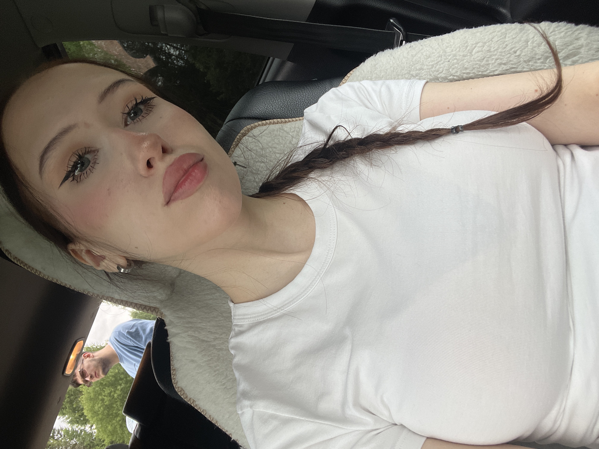 A young woman with long, dark brown hair braided down her back, wearing a white t-shirt, is sitting in the passenger seat of a car. She is looking at the camera with heavy eyeliner and light pink blush on her cheeks. Her lips are slightly parted and appear to be glossed. In the background, a man with short brown hair and glasses is visible in the driver's seat, looking out the window. He is wearing a light blue t-shirt. The interior of the car is visible, with a fluffy white sheepskin seat cover on the passenger side. The setting appears to be outdoors, with trees and greenery visible through the car windows, suggesting a rural or suburban environment. The location is identified as Gavrilovka, Russia.