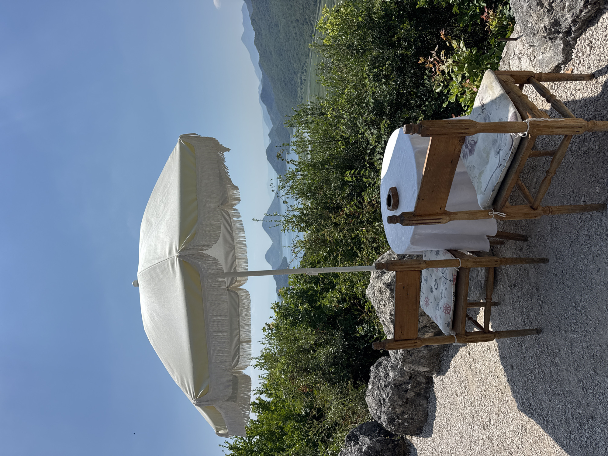 A white patio umbrella with fringe stands over a round table and two wooden chairs on a gravel patio. The table is covered with a white tablecloth and has an ashtray on it. The chairs have patterned seat cushions. In the background, a body of water is surrounded by green hills and a clear blue sky. The scene appears to be a scenic overlook or cafe with a view of a lake and mountains.
