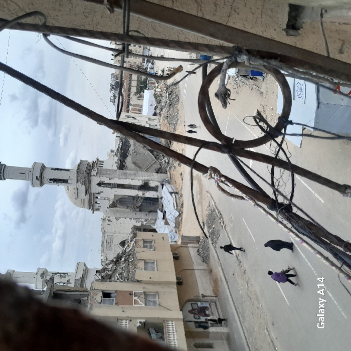 This image captures an urban scene in Khan Yunis, Palestinian Territory, from an elevated perspective, looking down onto a street and heavily damaged structures under an overcast sky.
The foreground is partially obscured by a dark, rusty metal railing and a tangle of wires. Below, a paved street with white lane markings forms the midground. On the street, a person in a purple top and dark trousers walks away from the camera, carrying a long-barreled weapon, likely a rifle, with both hands. Close by, another individual in a black full-body garment walks in the same direction. Further left, a third person in dark clothing walks across the street. In the distance, two more individuals are observed walking near a large debris pile.
To the left of the street, a multi-story building exhibits damage, including broken windows, exposed interiors, and hanging laundry on a balcony. A sign is visible on its ground floor. The background is dominated by a severely damaged mosque featuring two minarets and a central dome. Large sections of its walls have collapsed, exposing internal structures and creating extensive rubble mounds around its base, partially covered by white tarpaulins. Beyond the mosque, additional damaged buildings and debris are visible, emphasizing widespread destruction.