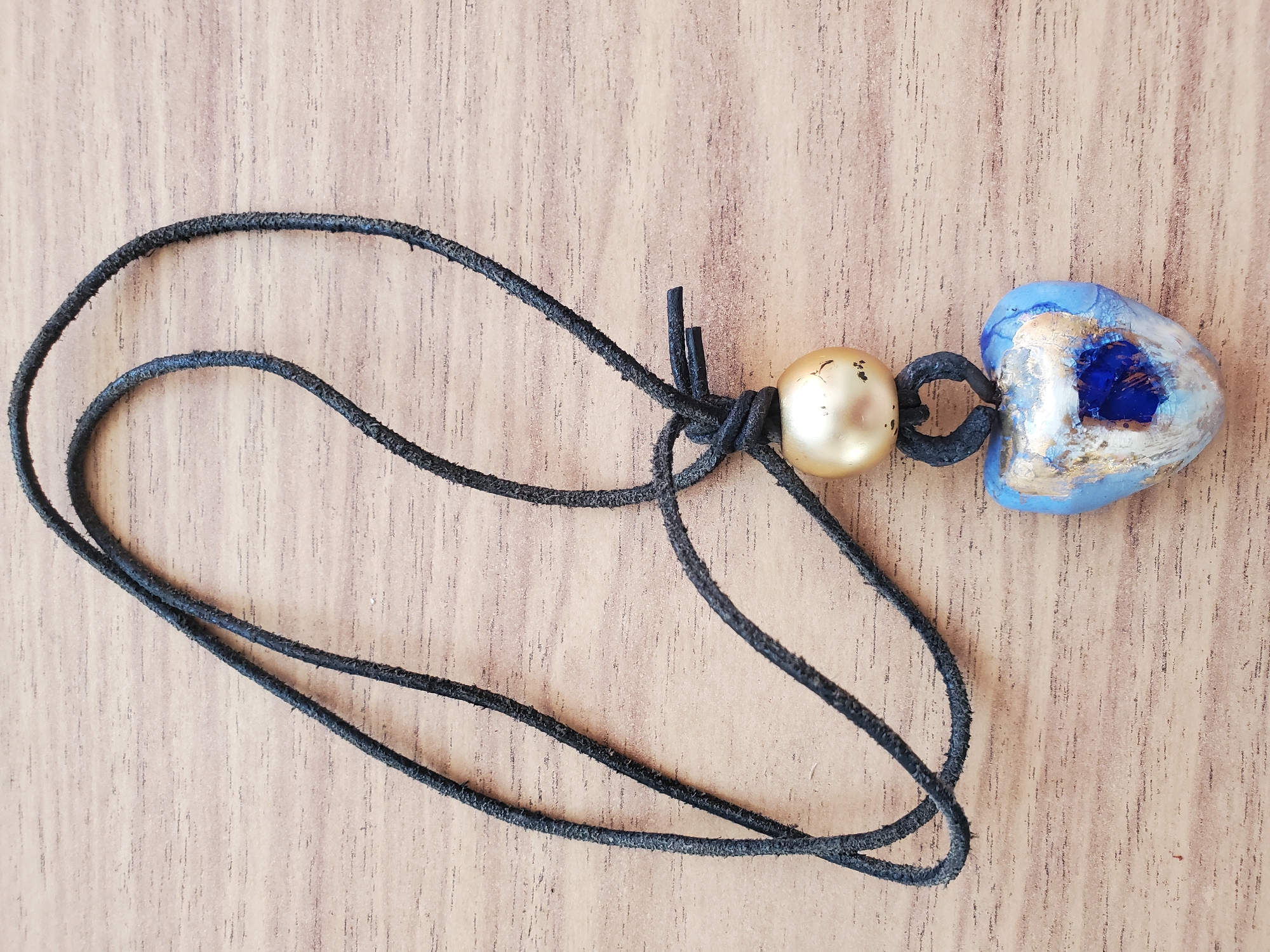 The image displays a close-up, overhead view of a handmade necklace resting on a wooden surface. The necklace consists of a black leather cord tied into a knot, from which hangs a gold-colored bead, a small black loop, and a heart-shaped pendant. The pendant is primarily blue with streaks of gold and a central darker blue, textured element. The wooden surface exhibits a prominent grain pattern. There is no indication of the city of Tiraspol, Moldova, or any people or actions within the image.